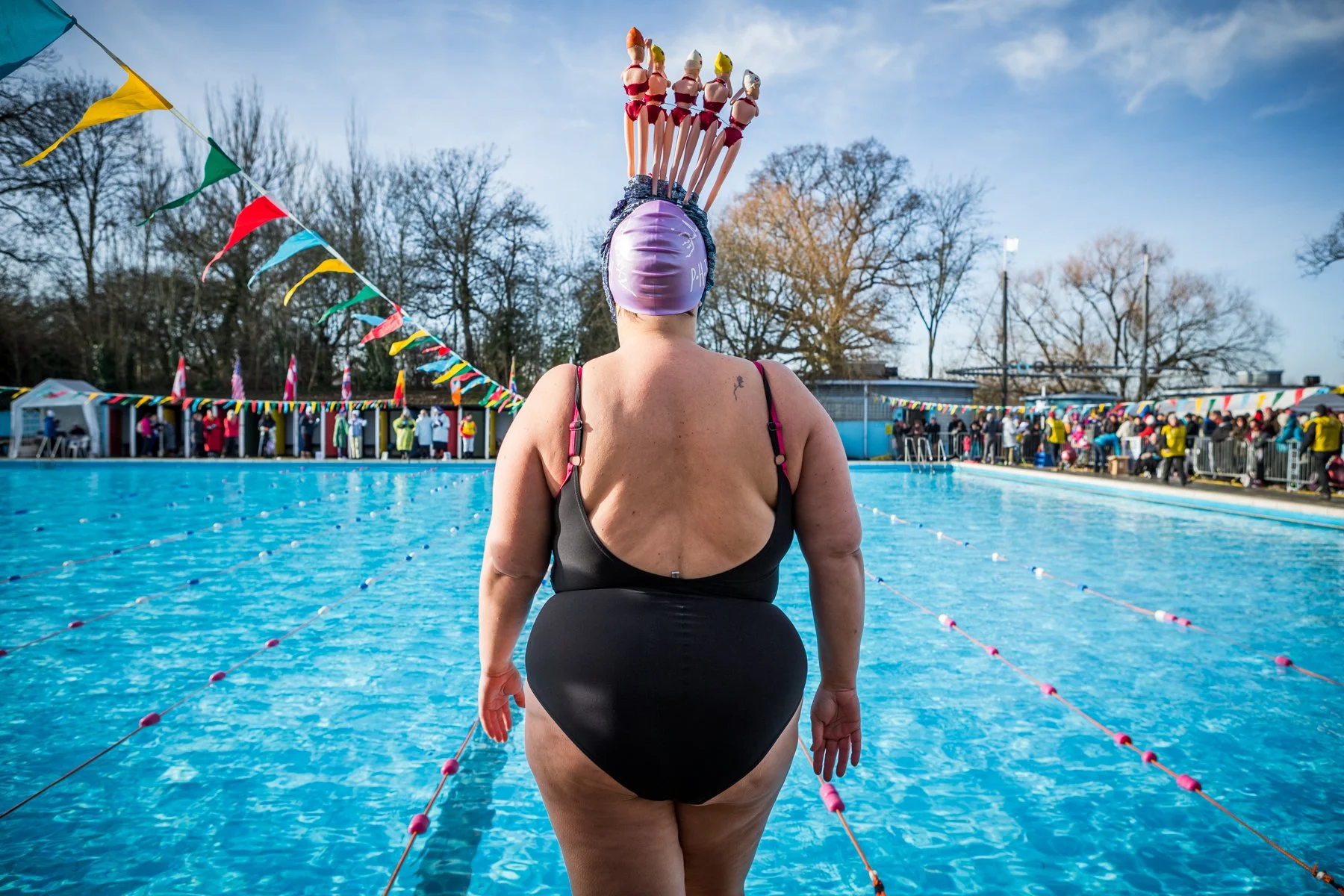 A woman standing at the edge of an outdoor swimming pool, wearing a black swimsuit and a purple swim cap with a floatation device on top, while a group of people in colorful clothing and a row of small flags are visible in the background.