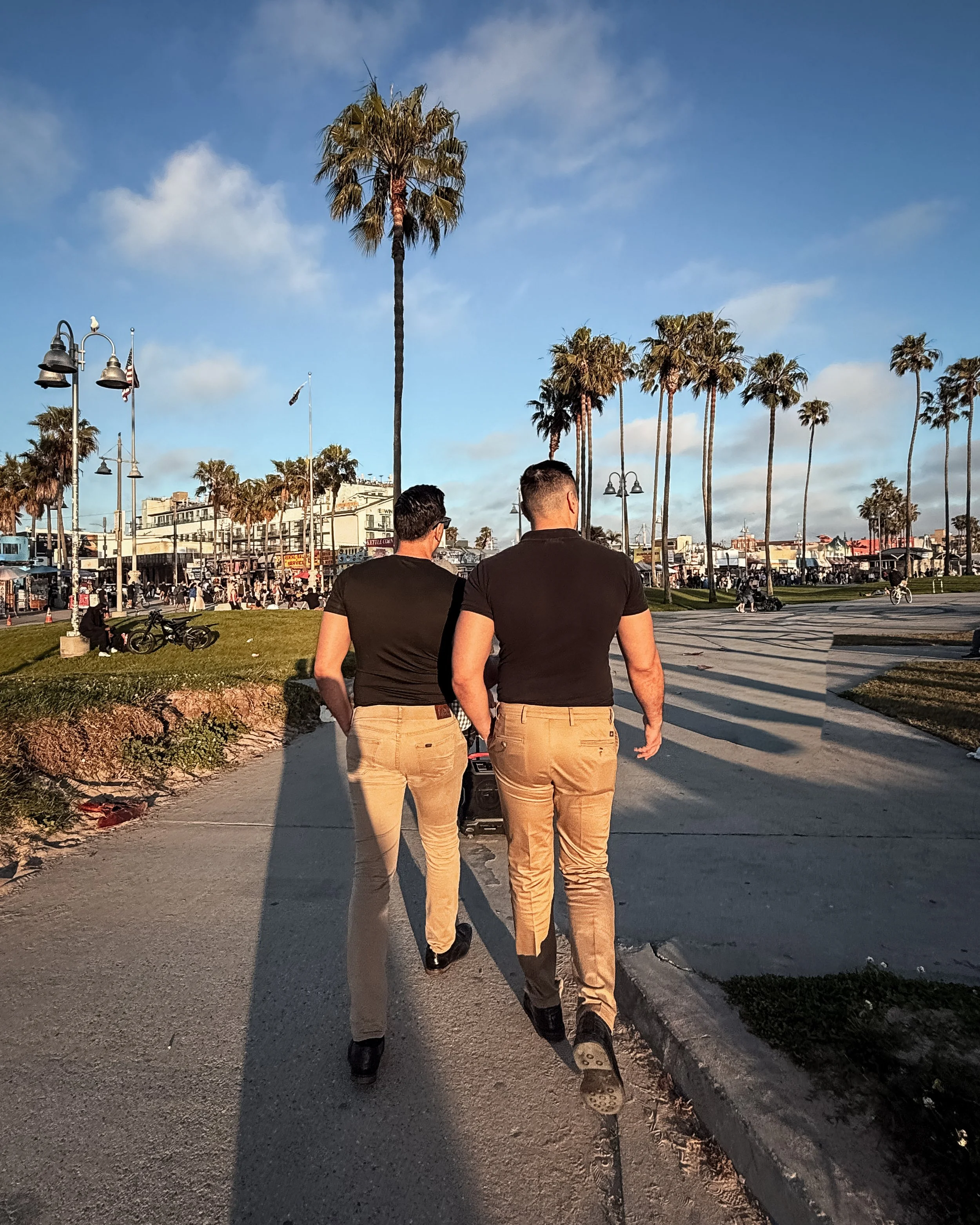 Two men walk down a sidewalk in a sunny area with palm trees, buildings, and a crowd in the background.