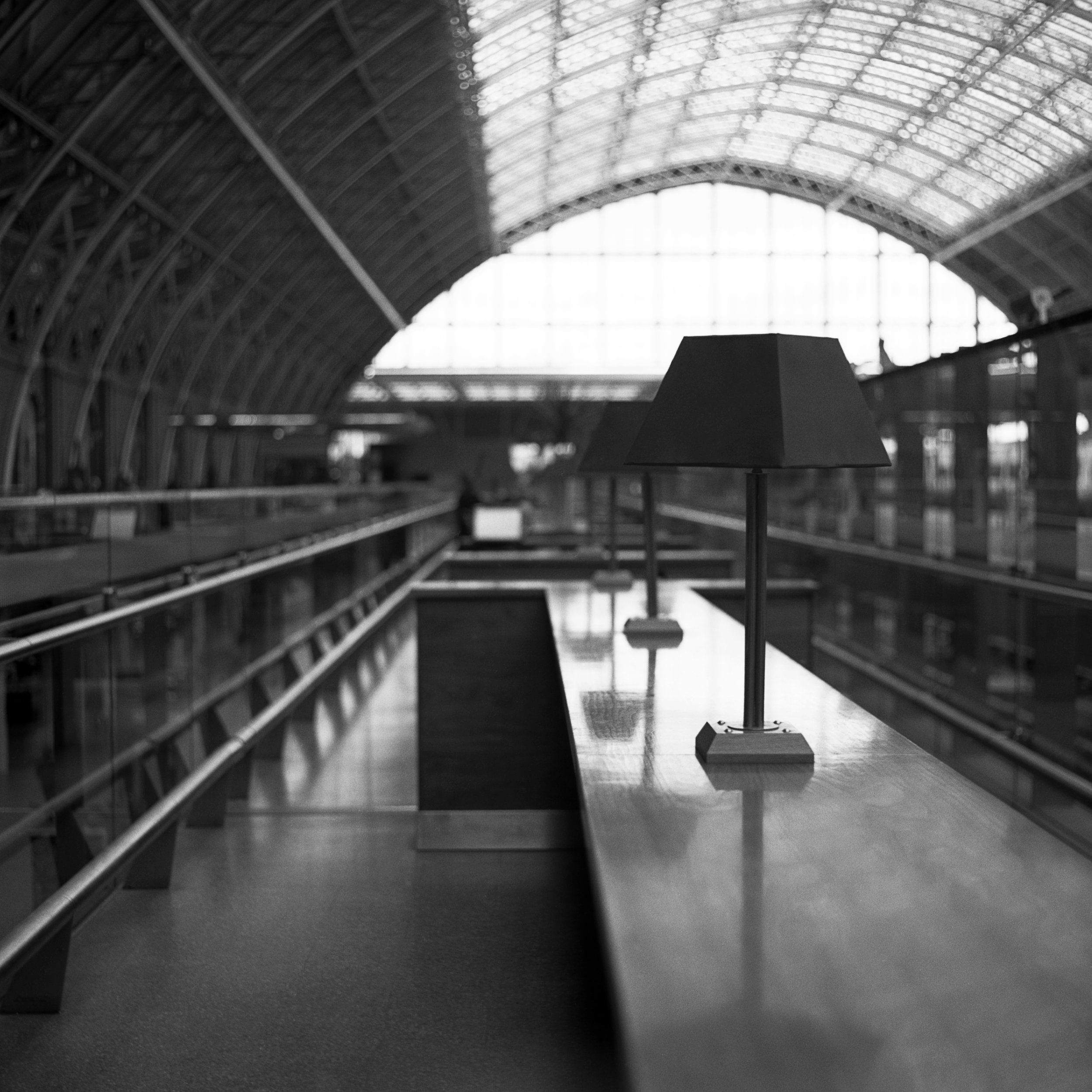 Black and white photo of an indoor train station or airport terminal with high arched ceiling and large windows, featuring a solitary lamp on a tall stand in the foreground.