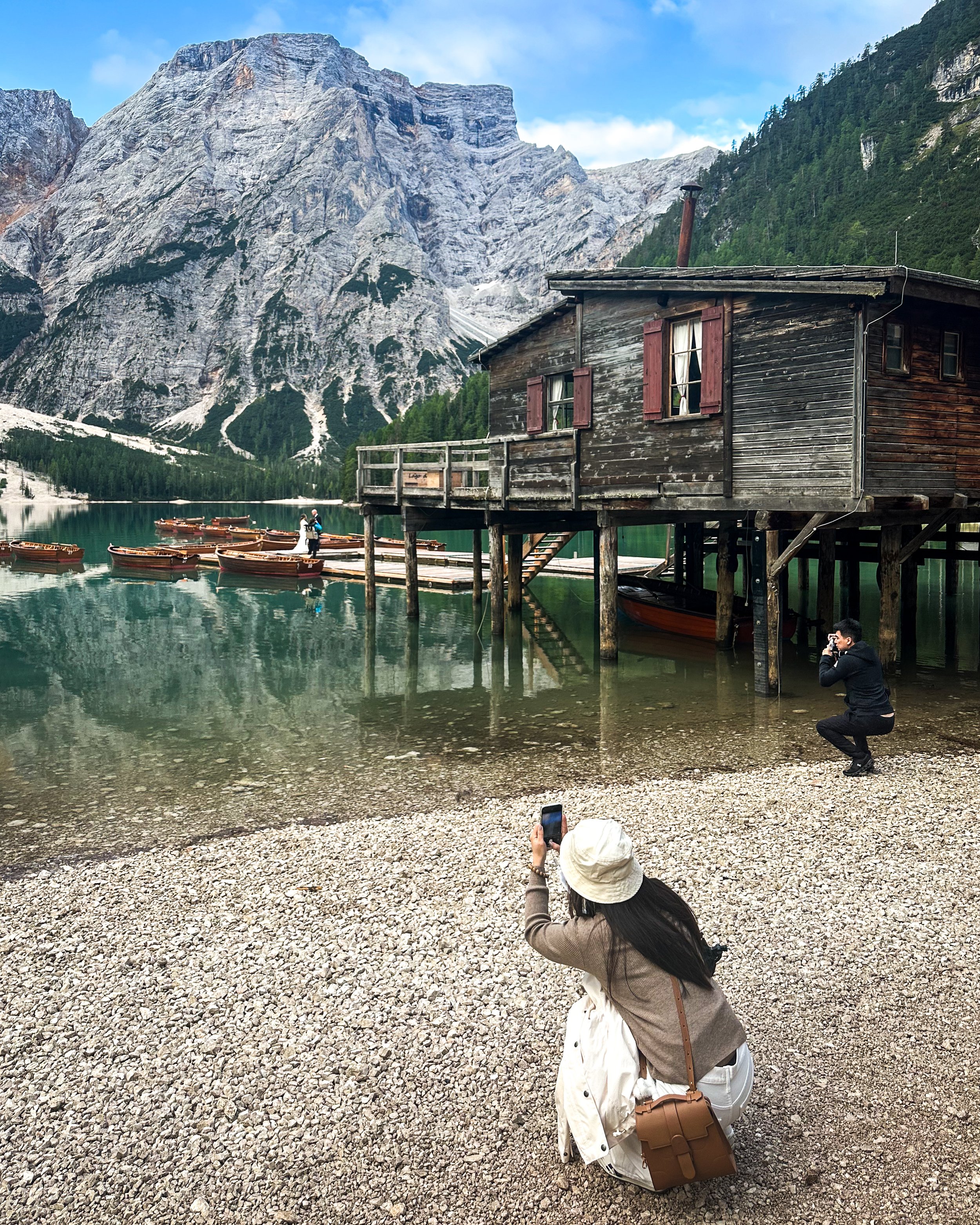 People taking photos of a wooden cabin on stilts by a lake, with a large mountain range and green forest in the background.
