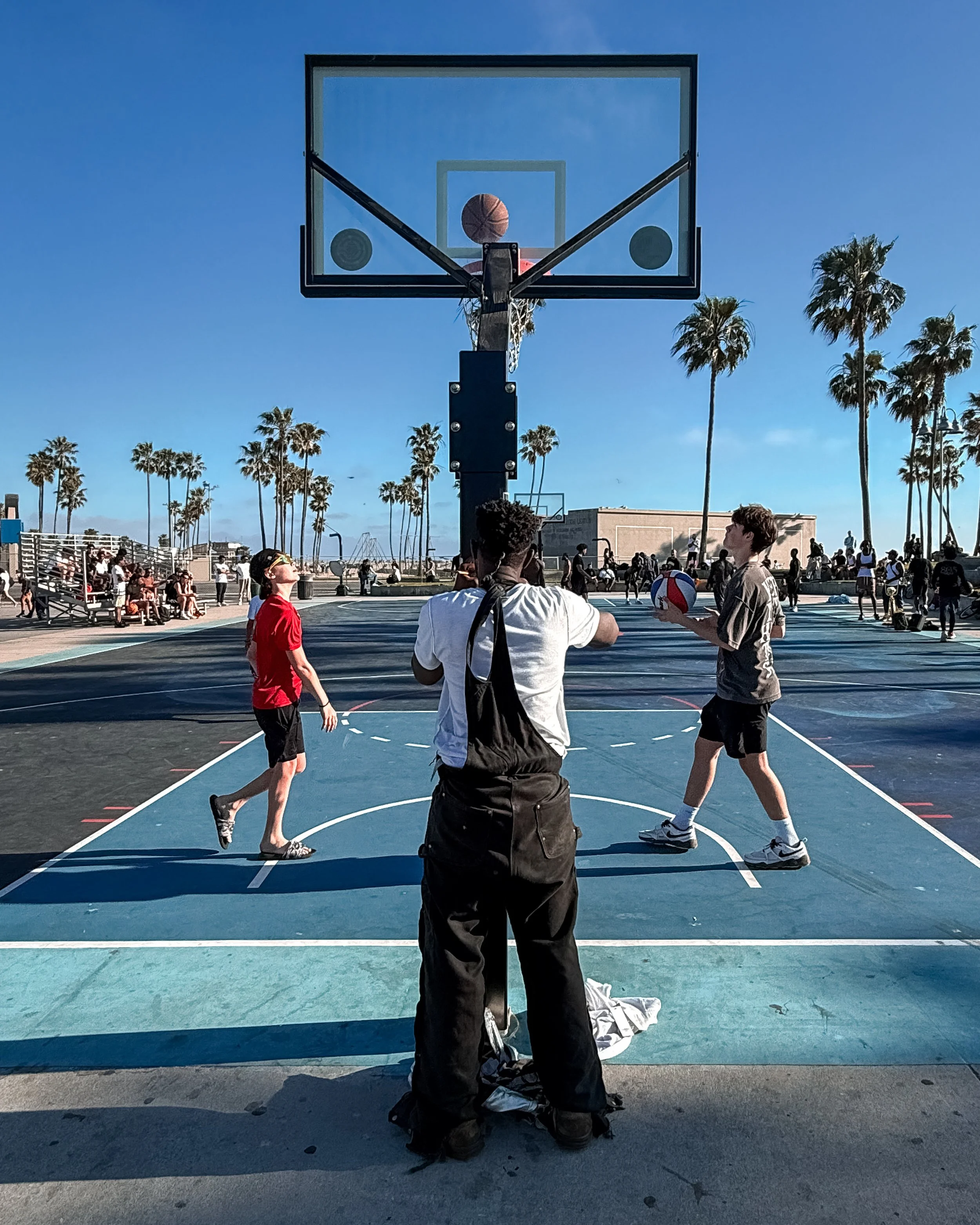 A group of young people playing basketball on an outdoor court under a clear blue sky, with palm trees and spectators in the background.