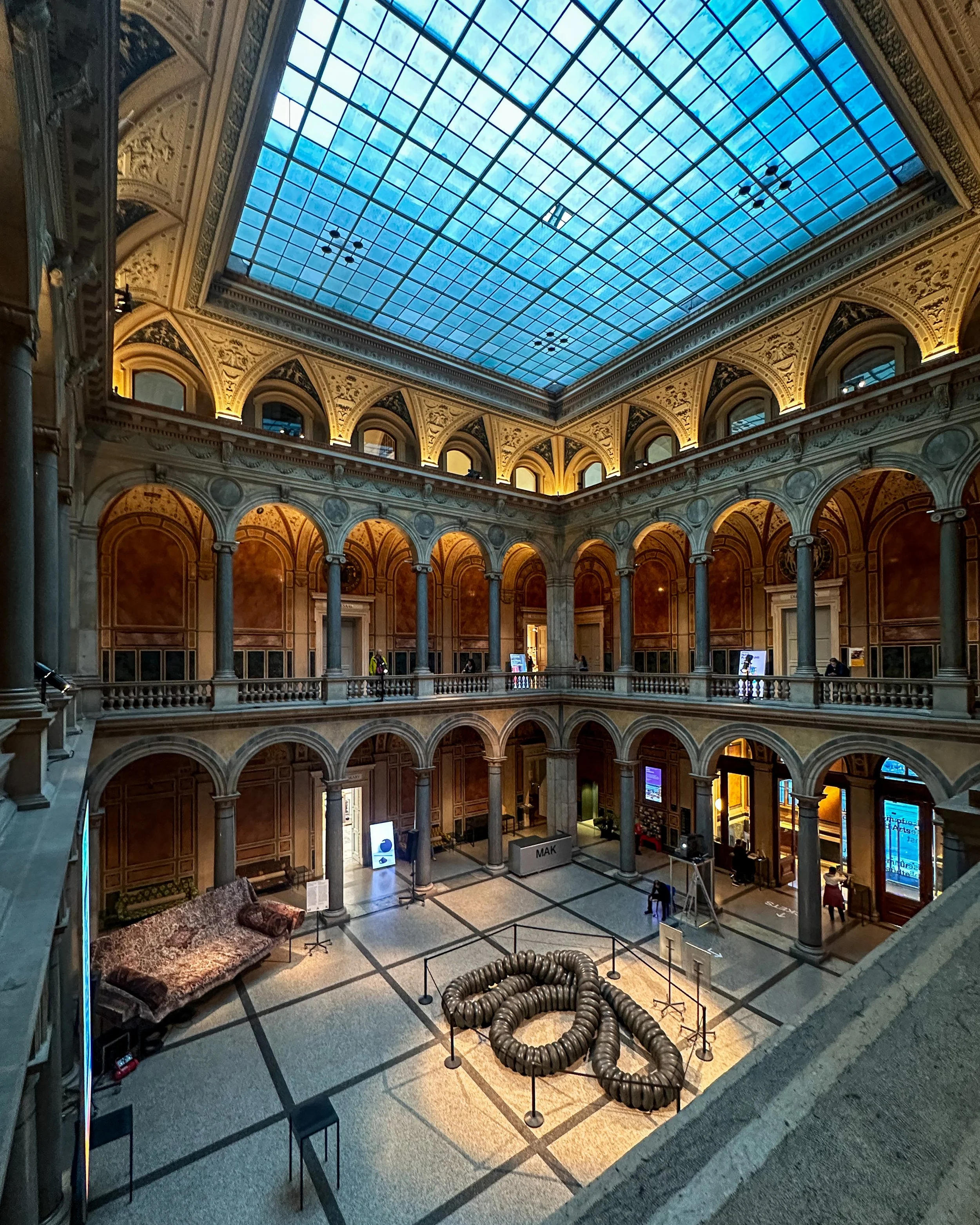 Interior view of a historic building with a large glass roof, arches, columns, and decorative interior details. The ground floor has an art installation composed of large metal coils arranged in a spiral shape, surrounded by barriers, with a vintage 
