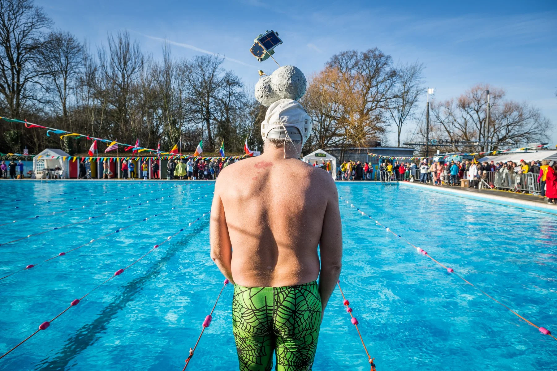 A swimmer standing at the edge of an outdoor swimming pool, wearing green patterned swim trunks and a white swim cap with a large rock on top that holds a small model space shuttle and satellite. There are spectators and booths in the background, wit