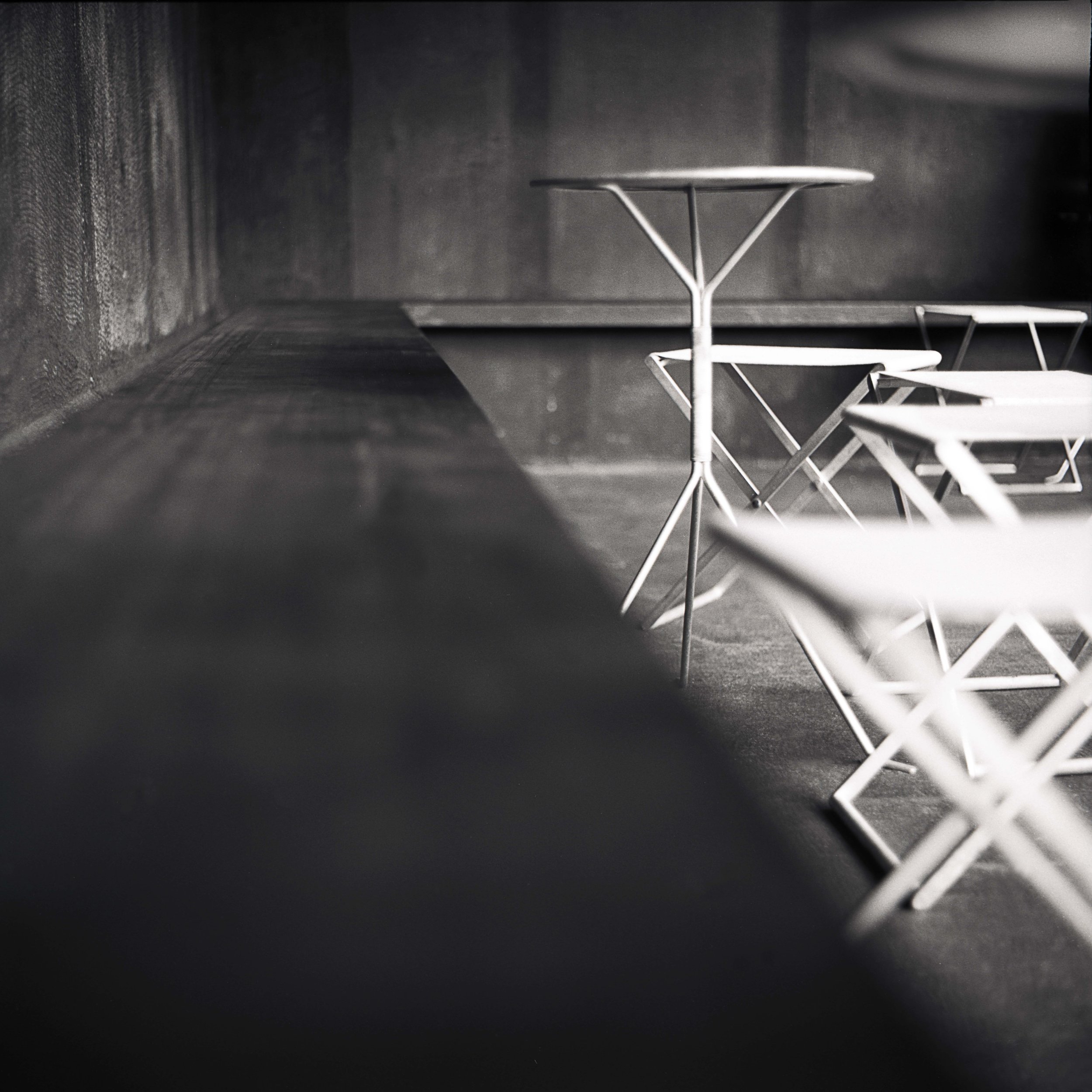 A black and white photo of a minimalistic wire chair with a round seat and a matching wire side table, positioned against a dark wall at the Serpentine Pavilion in London, United Kingdom.