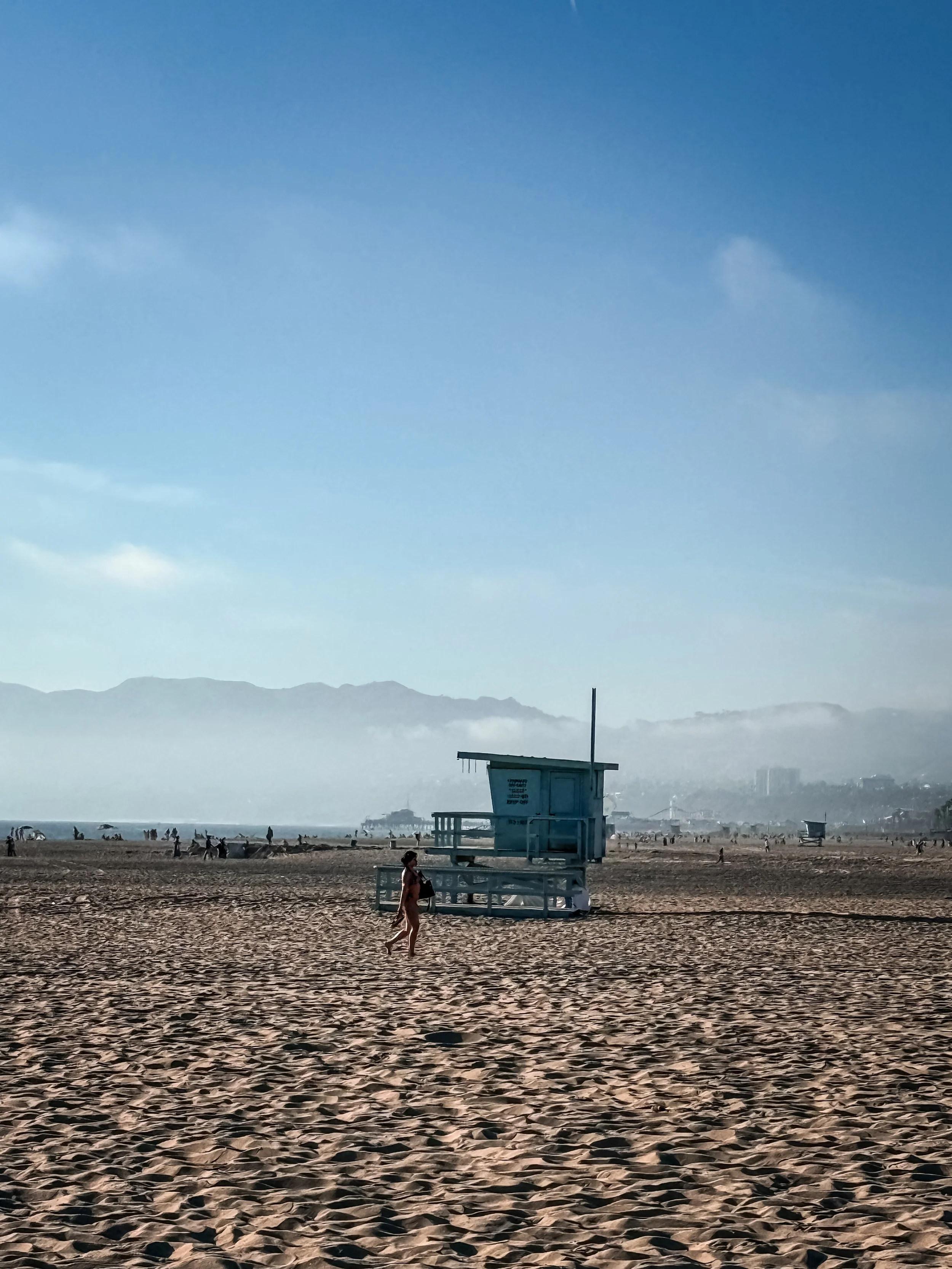 Beach scene with a lifeguard tower, people walking and relaxing, distant mountains, and a hazy sky.