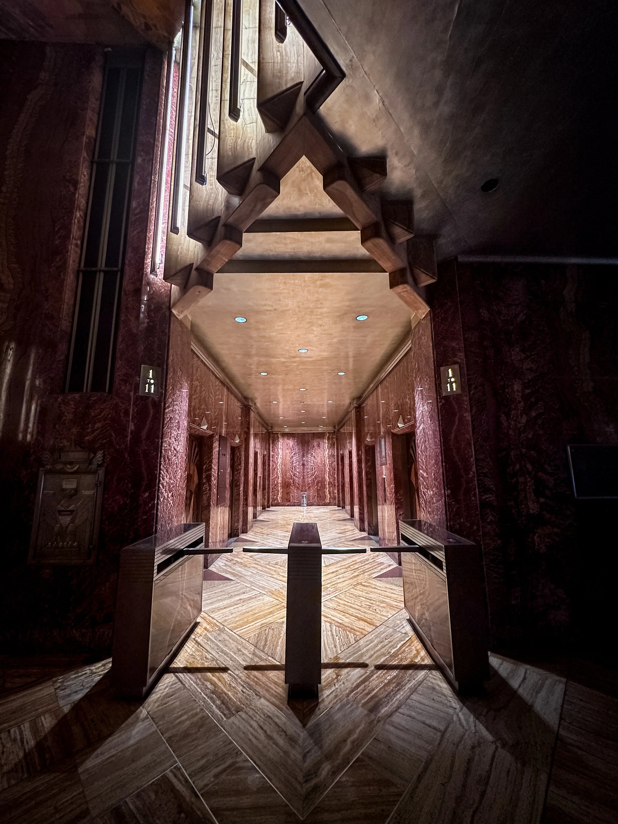 An interior hallway with maroon marble walls, a wooden ceiling, and a patterned marble floor. There are elevator doors on both sides with gold directional signs, and a glass display case on the left. The ceiling has built-in lights.
