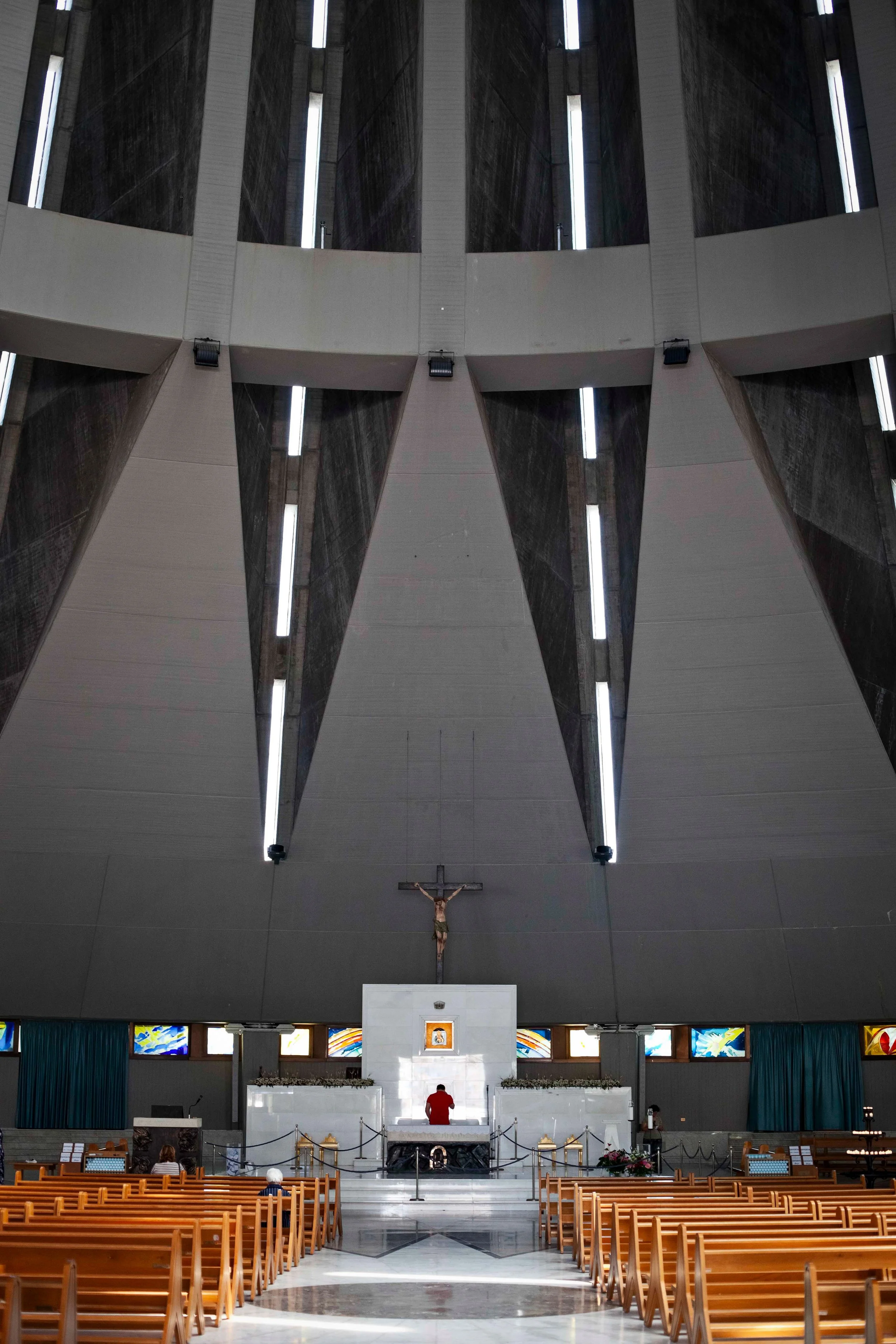 Interior of the Basilica of the Madonna delle Lacrime, a Roman Catholic church in Syracuse in Sicily, Italy. Wooden pews face an altar, with a person standing at the altar and a crucifix hanging above.