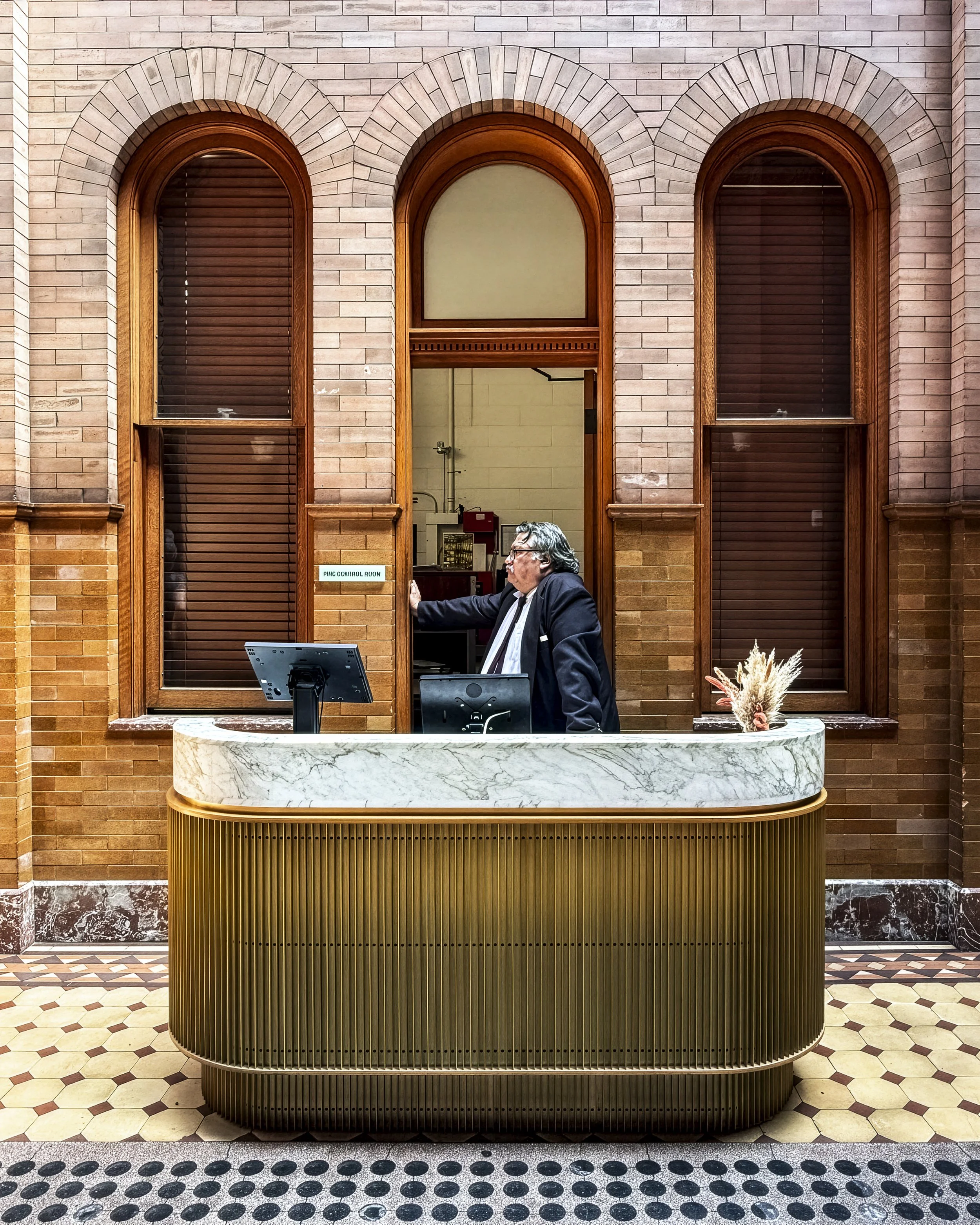 Interior view of the Bradbury Building in Downton Los Angeles, California, USA. A man in a dark suit stands behind a marble reception desk in front of a brick wall with three arched windows.
