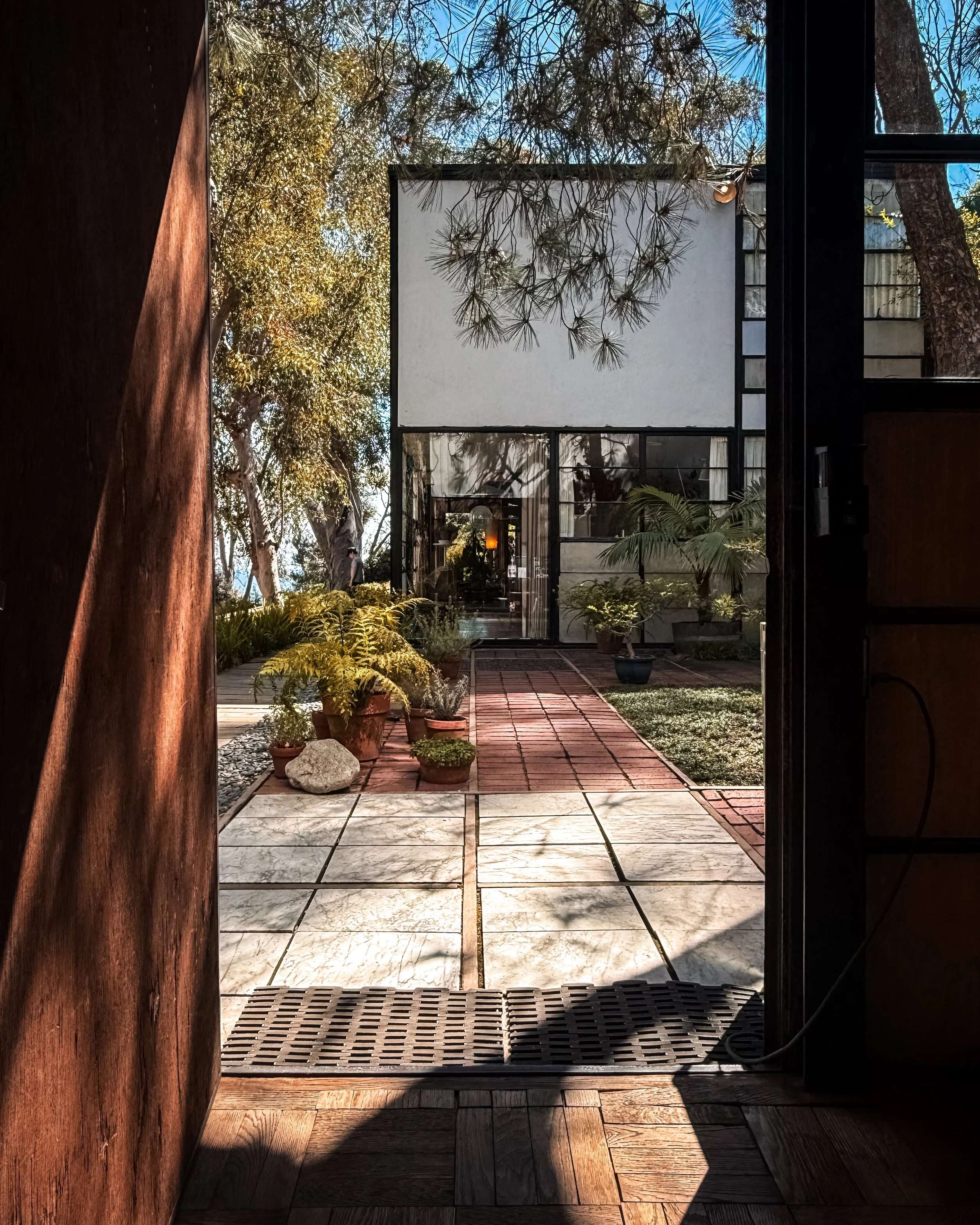 Charles and Ray Eames' Eames House in Los Angeles, California, USA, part of the Case Study House Program. View from inside the kit looking out through a door at a patio area with potted plants, brick tiles, and trees. Architecture photography.