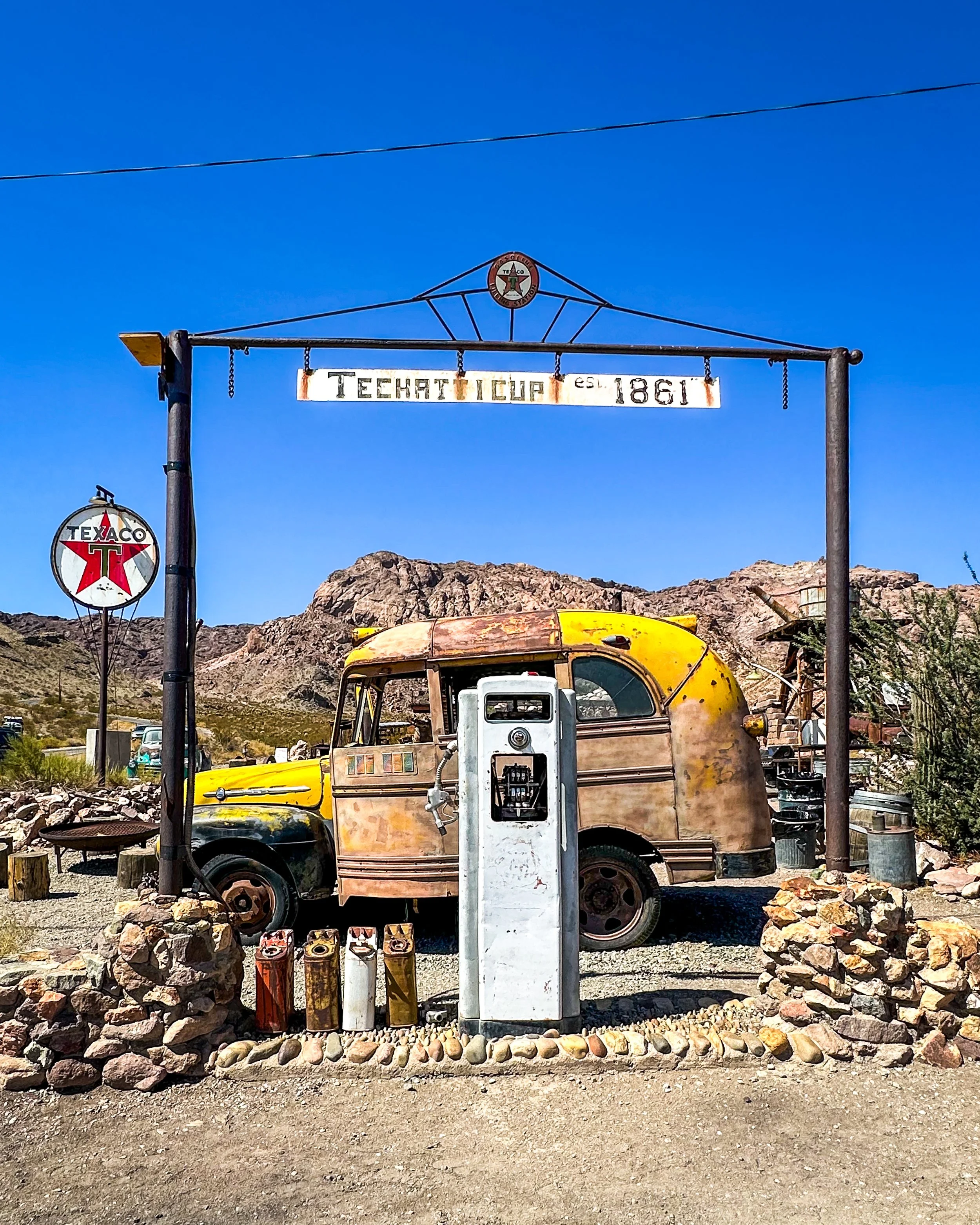 Vintage gas pump with a rusted yellow vehicle behind it, surrounded by rocks and stones, with an old Texas Company sign and a weathered sign that says 'TECHATTICUP EST. 1861' against a mountainous landscape under a clear blue sky.
