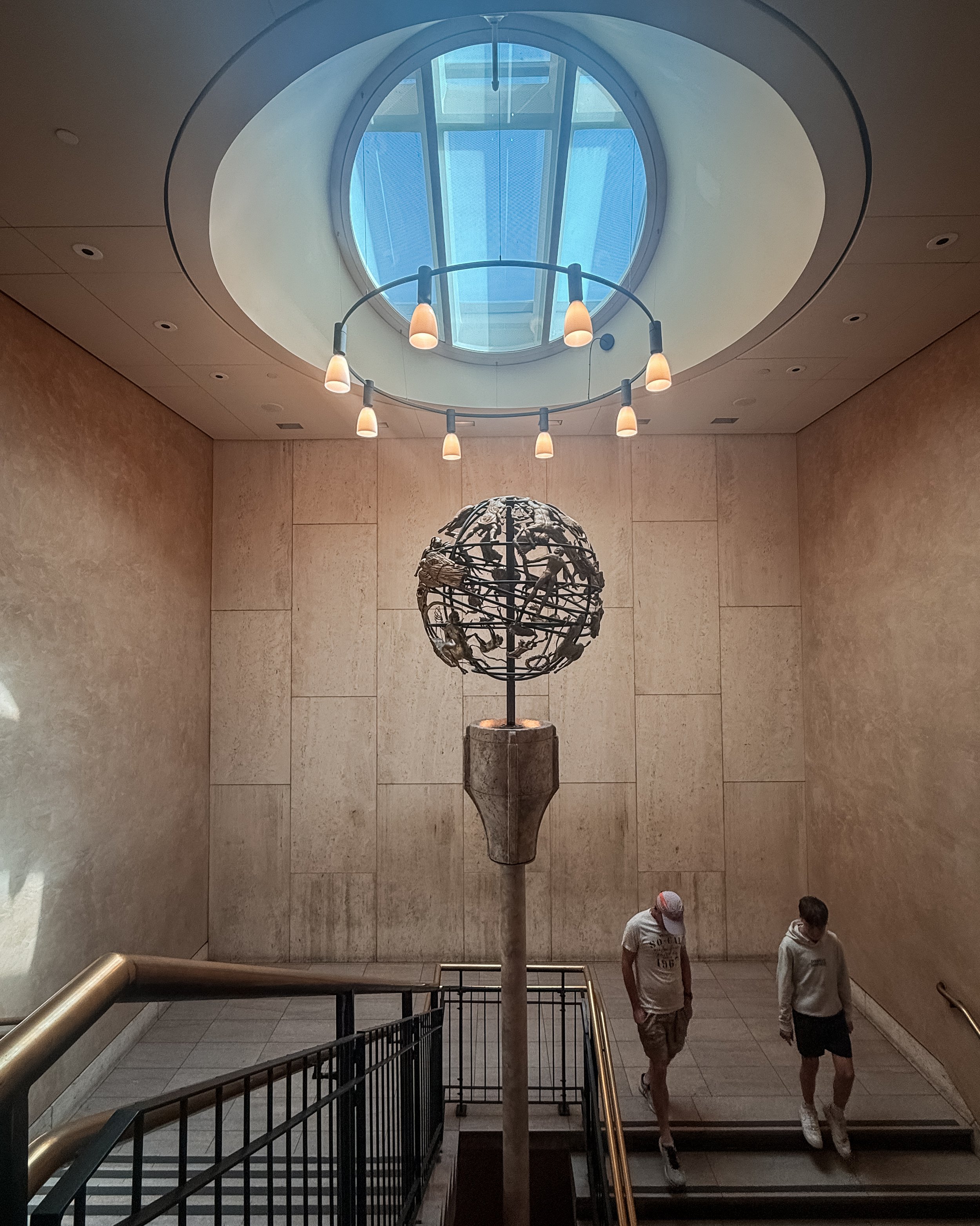 Interior view of a stairwell with a large circular art piece resembling a globe, two people walking down the stairs, and a large skylight on the ceiling letting in natural light.