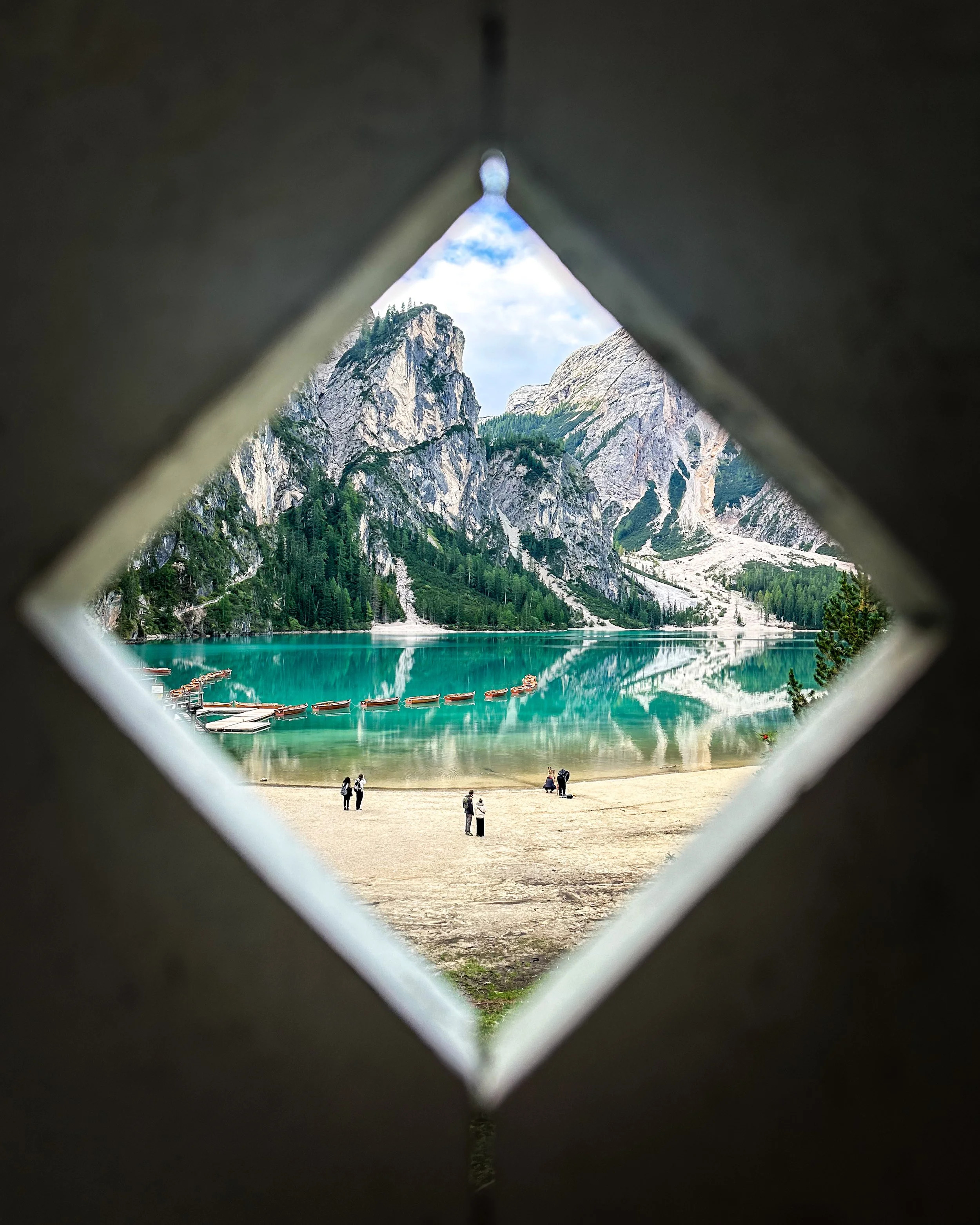 View through a diamond-shaped opening in a metal fence shows a lake with docked boats, surrounded by rocky mountains and green forests, with people walking along the shore.