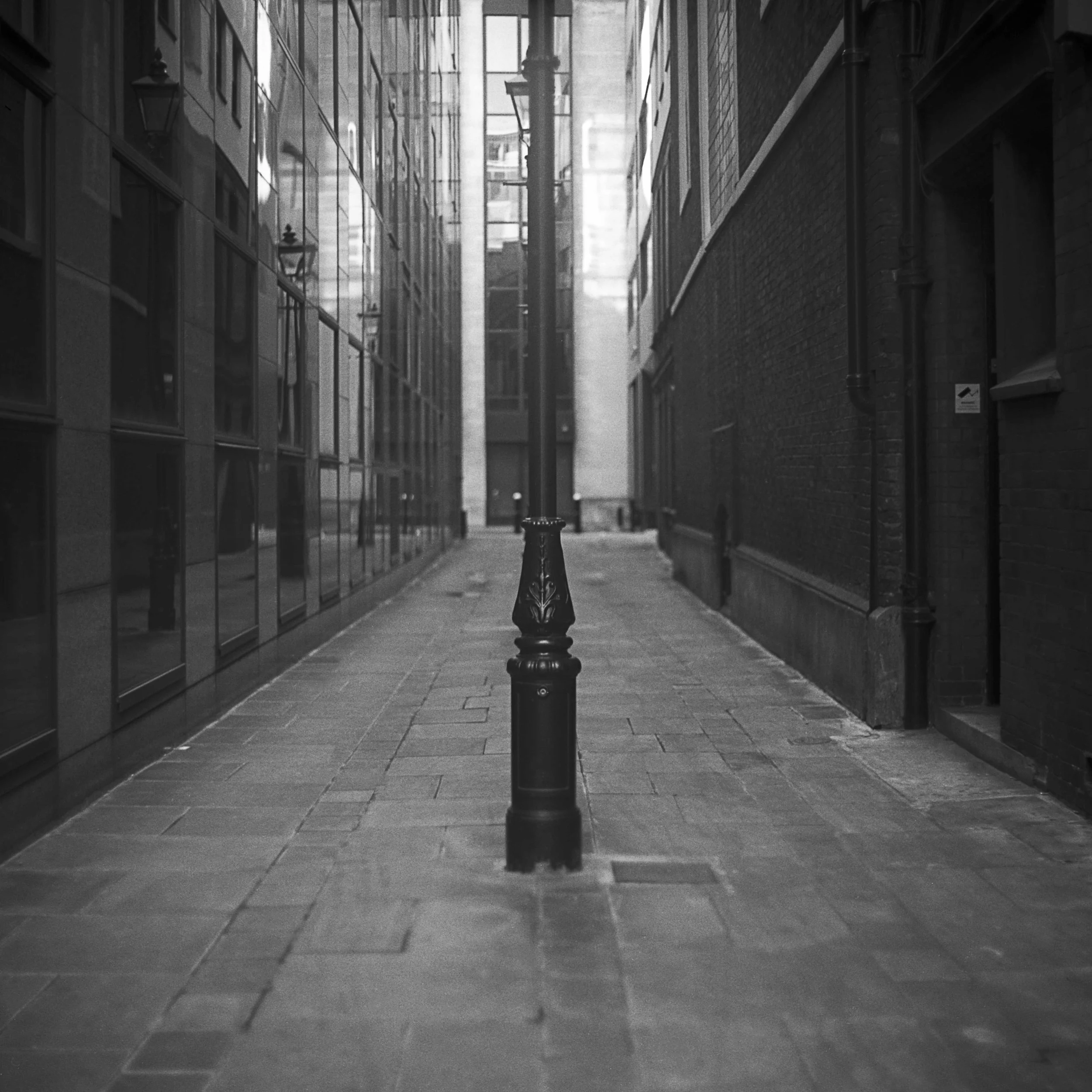 A black and white film photo of a narrow urban alleyway in London, United Kingdom, flanked by tall buildings, with a decorative bollard in the foreground.
