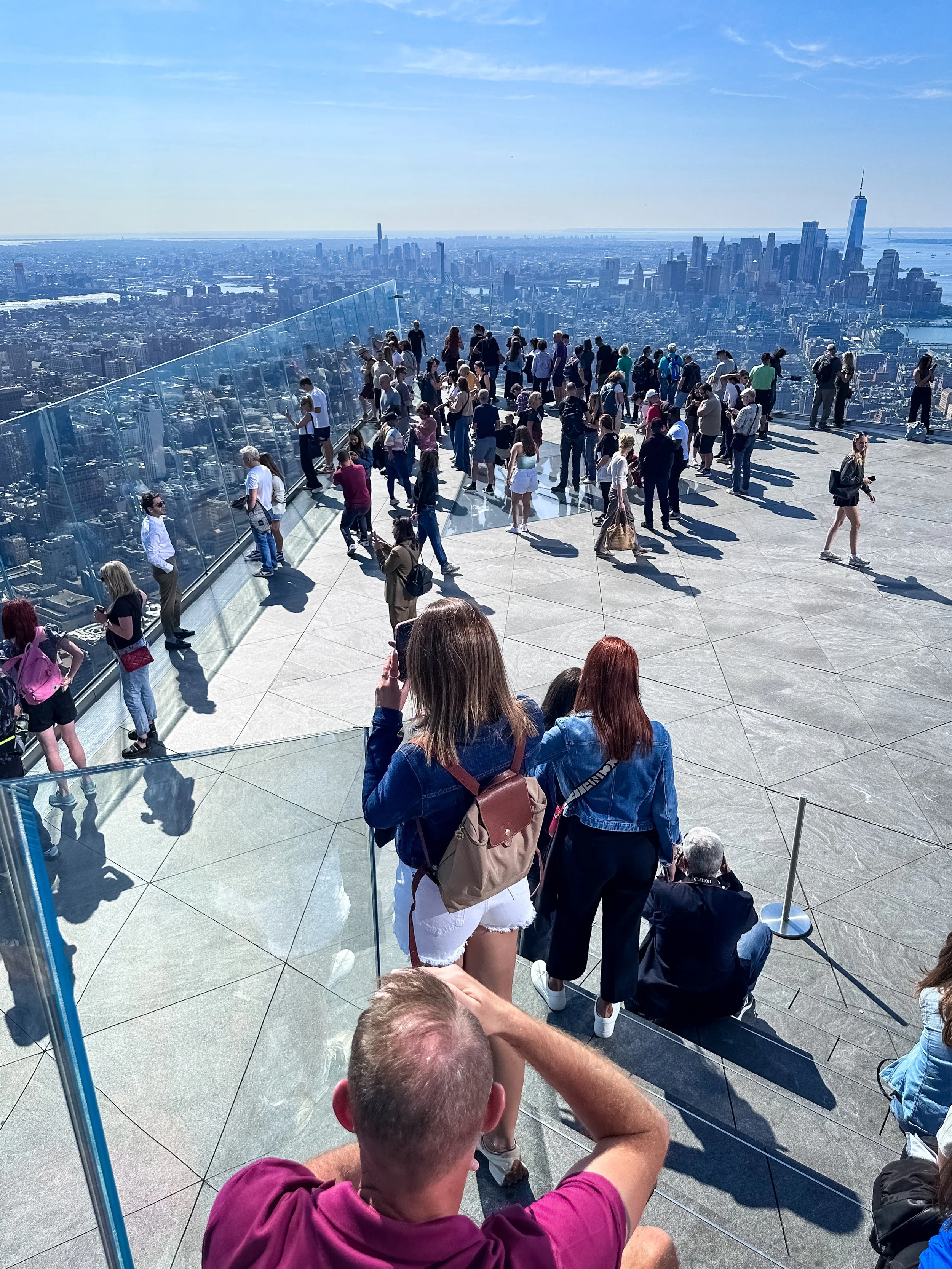 People standing and sitting on an observation deck overlooking the New York City skyline with tall skyscrapers, including One World Trade Center, and the East River in the background.