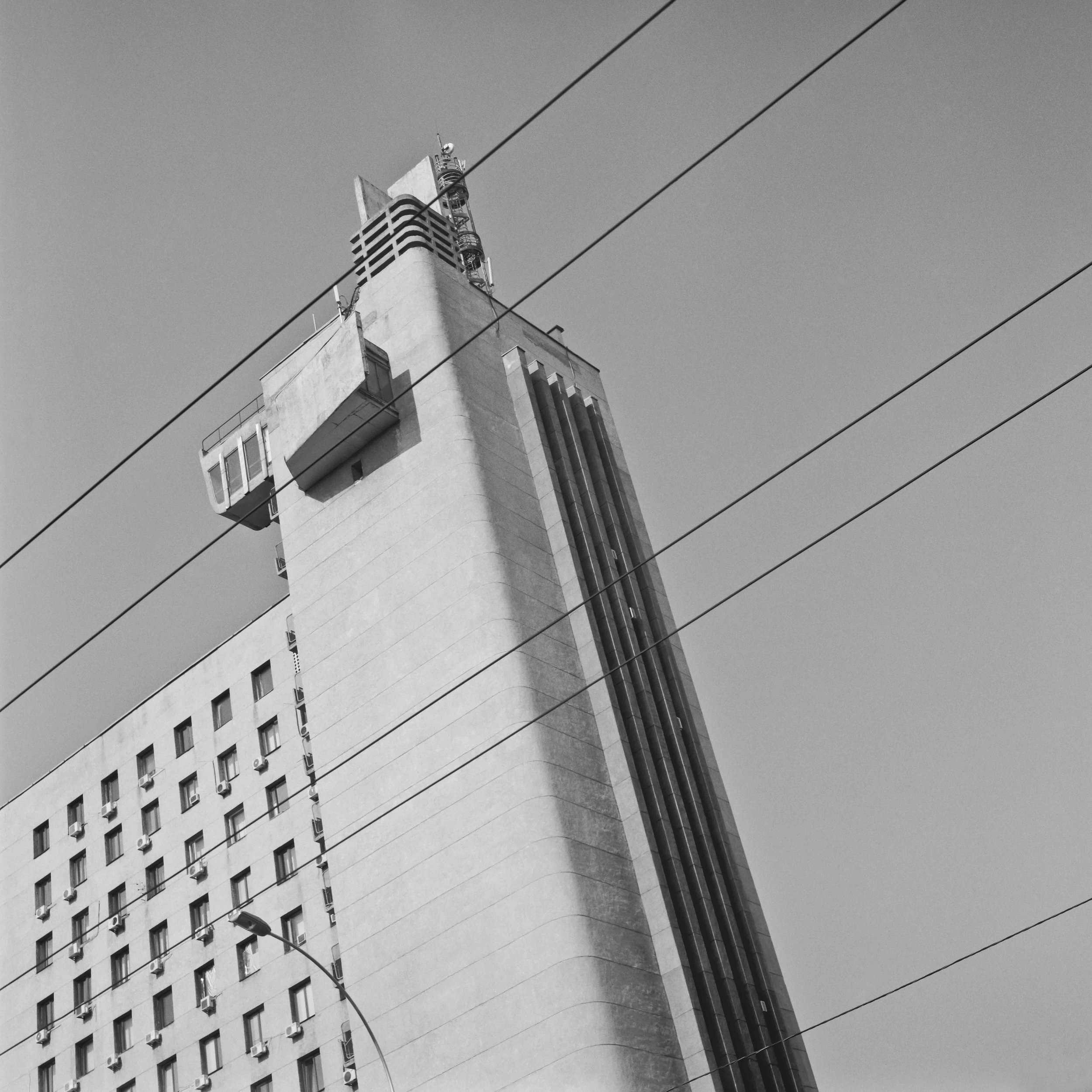 A black-and-white photo of the top part of a tall building with multiple windows and a protruding structure at the rooftop, with four power lines crossing the image diagonally. Black and white architecture photograph shot in Kyiv, Ukraine.