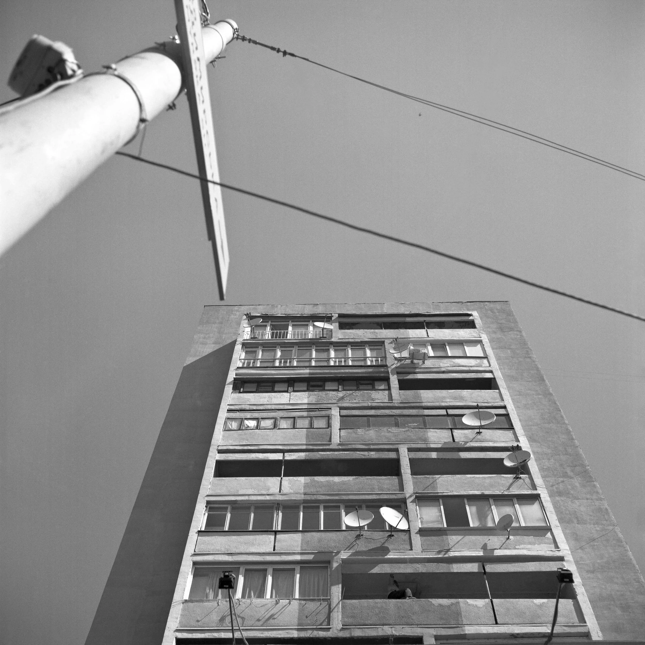 Looking up at a tall multi-story building with satellite dishes and antennas attached to the balcony railings, and cables extending into the sky. Black and white architecture photograph shot in Kyiv, Ukraine.