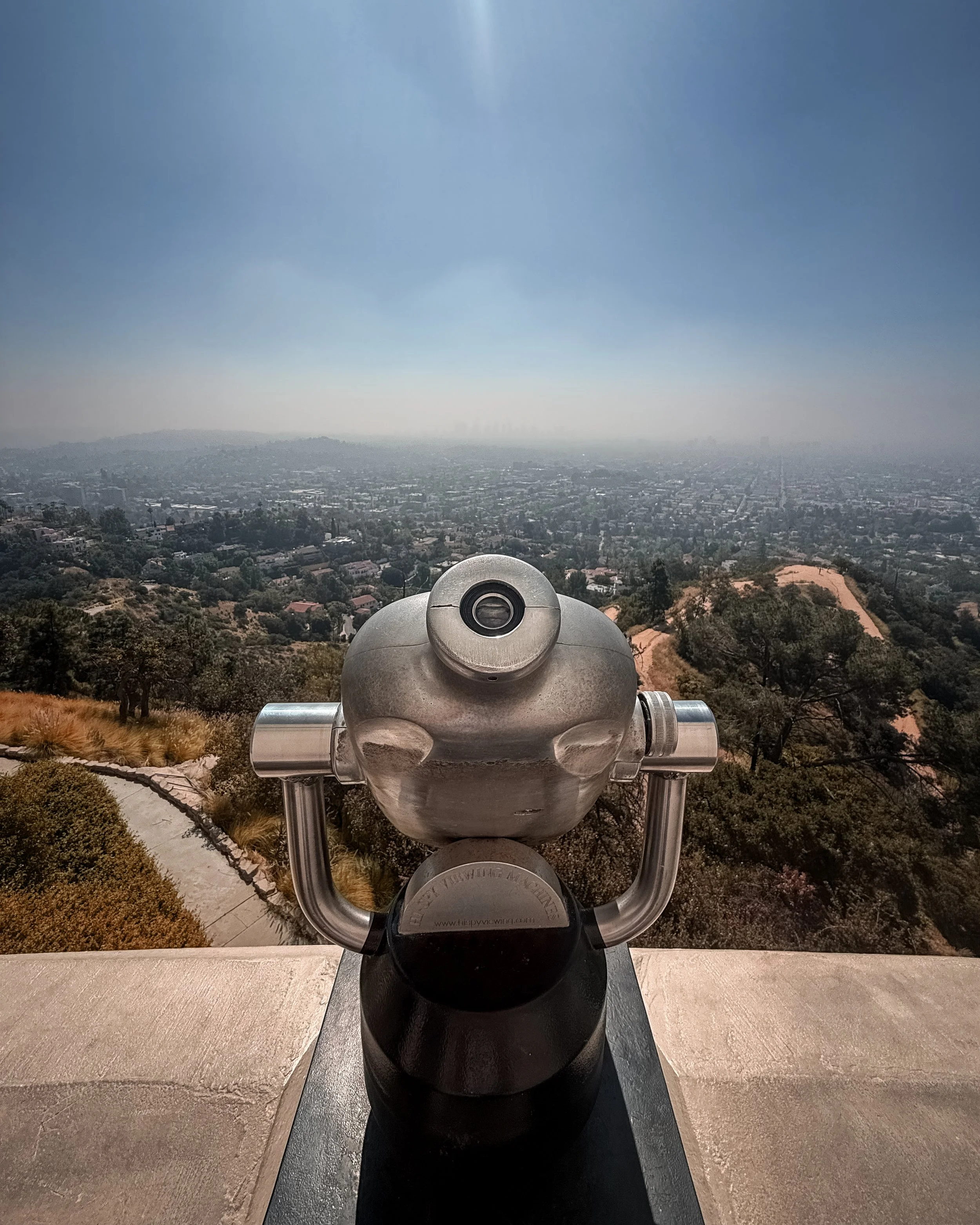 A view from a hilltop overlooking a cityscape with a coin-operated viewing telescope in the foreground, clear sky above, and distant city skyline.