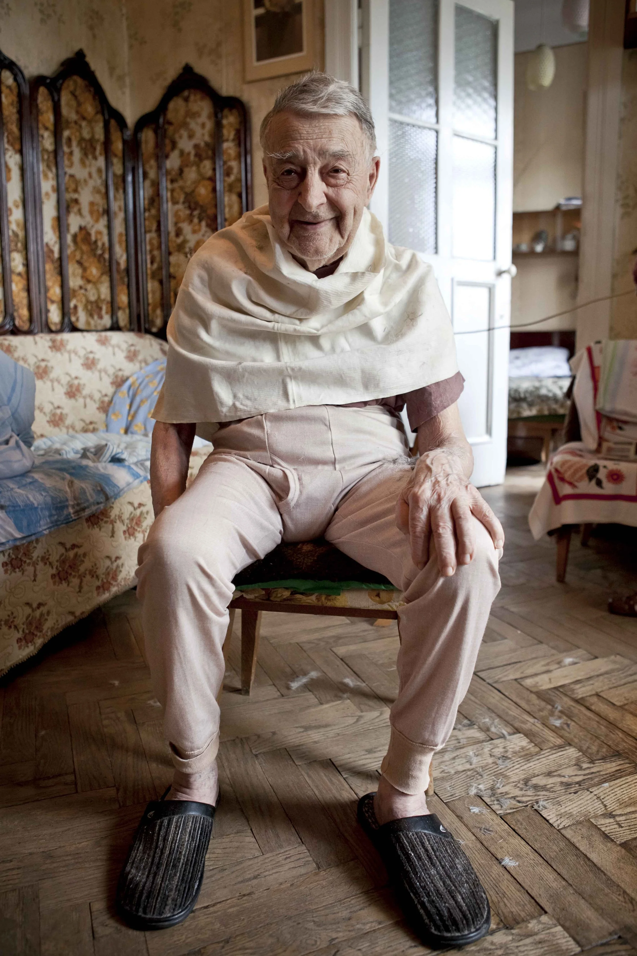 An elderly man sitting on a wooden chair smiling at the camera in Kyiv, Ukraine. He is about to have his hair cut, so has cream-coloured drape around his shoulders. 