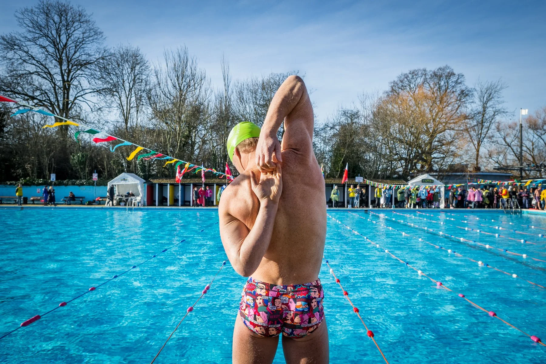 A male swimmer wearing a green swim cap and colorful swim trunks with comic book style graphics, stretching and adjusting his swim cap by the poolside during a swimming event. In the background, there are spectators and other swimmers, with trees and
