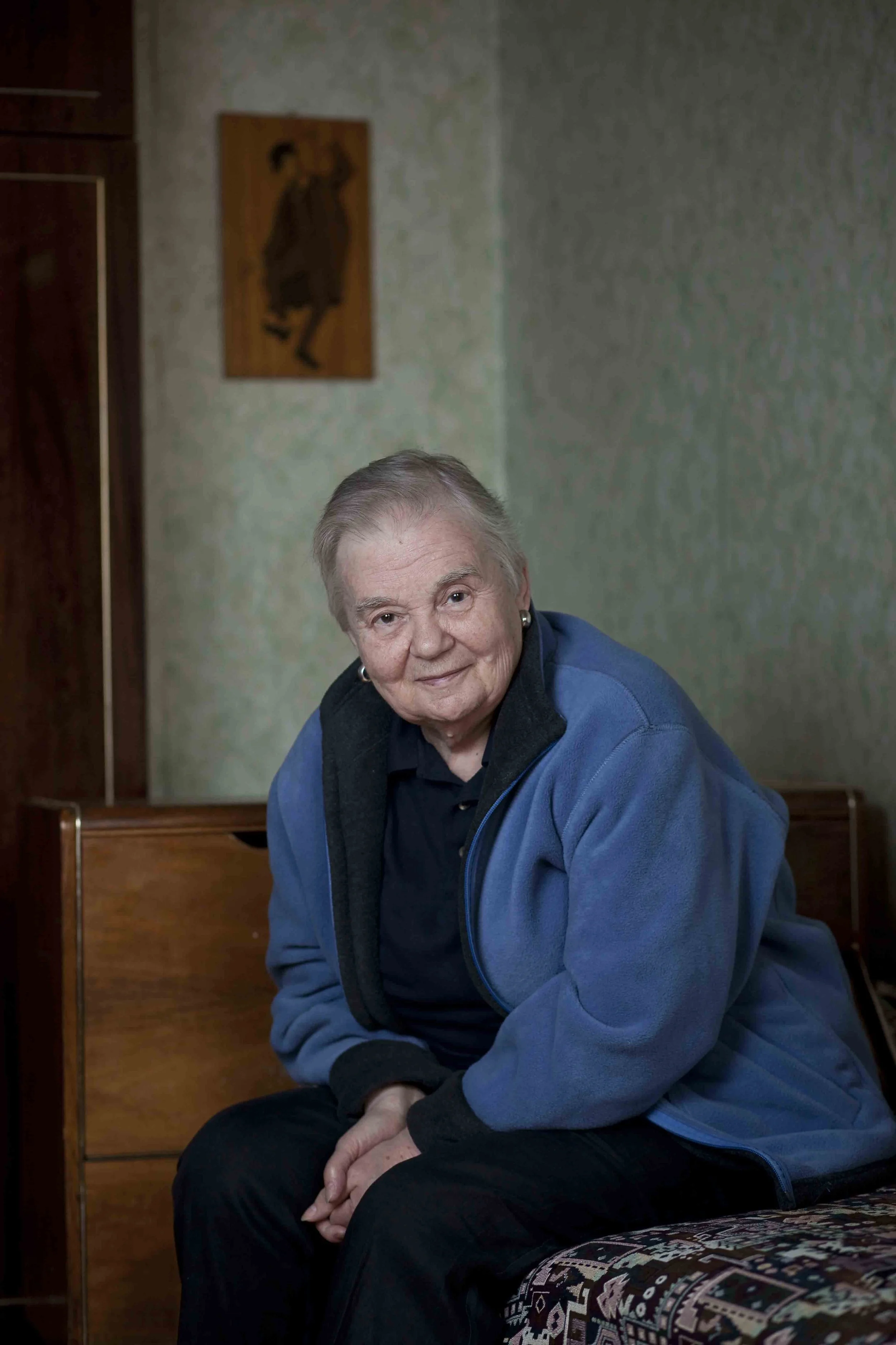 An elderly woman with short gray hair, wearing a black and blue jacket, sits on a chair in a room with green patterned walls. There is a wooden cabinet on her left and a framed abstract art piece on the wall behind her.