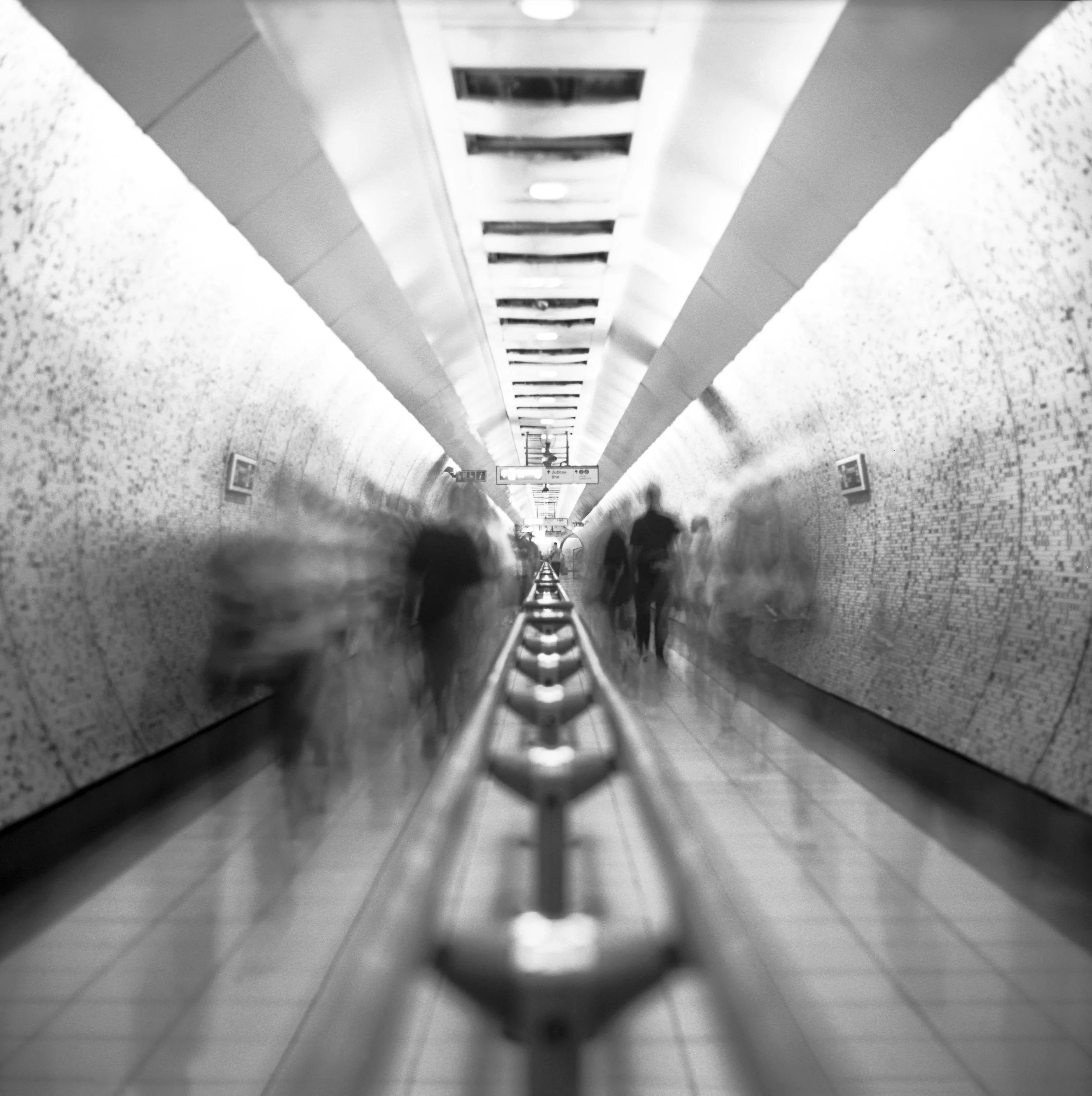 Looking along a subway or tube station corridor with tiled walls and a row of lights on the ceiling, partially blurred, with silhouettes of people in motion. London, United Kingdom.