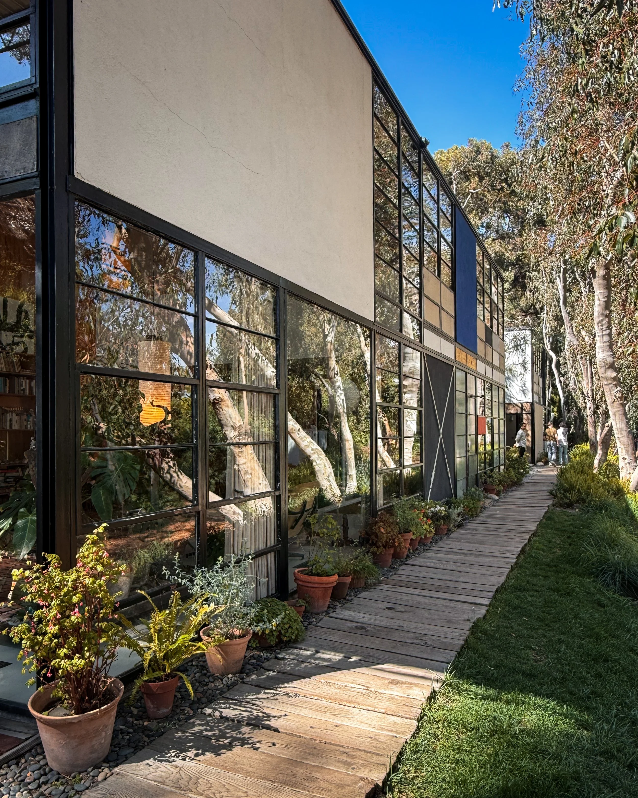 Charles and Ray Eames' Eames House in Los Angeles, California, USA, part of the Case Study House Program. The house is surrounded by potted plants and trees, with a wooden pathway and green grass in the foreground. Architecture photography. 