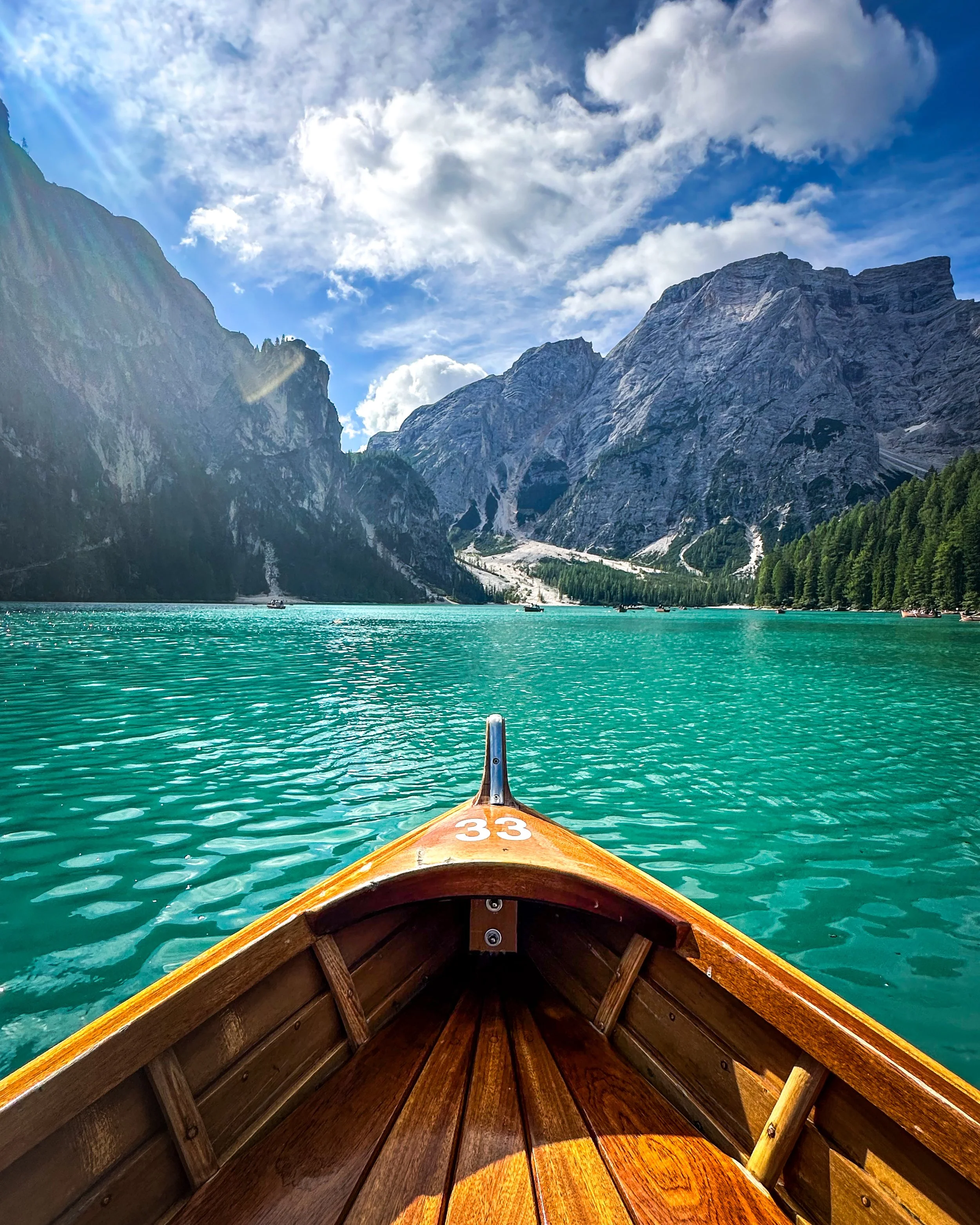View from a canoe on a lake (Lago di Braies - Pragser Wildsee in the Dolomites, Italy) surrounded by mountains with trees, under a partly cloudy blue sky.