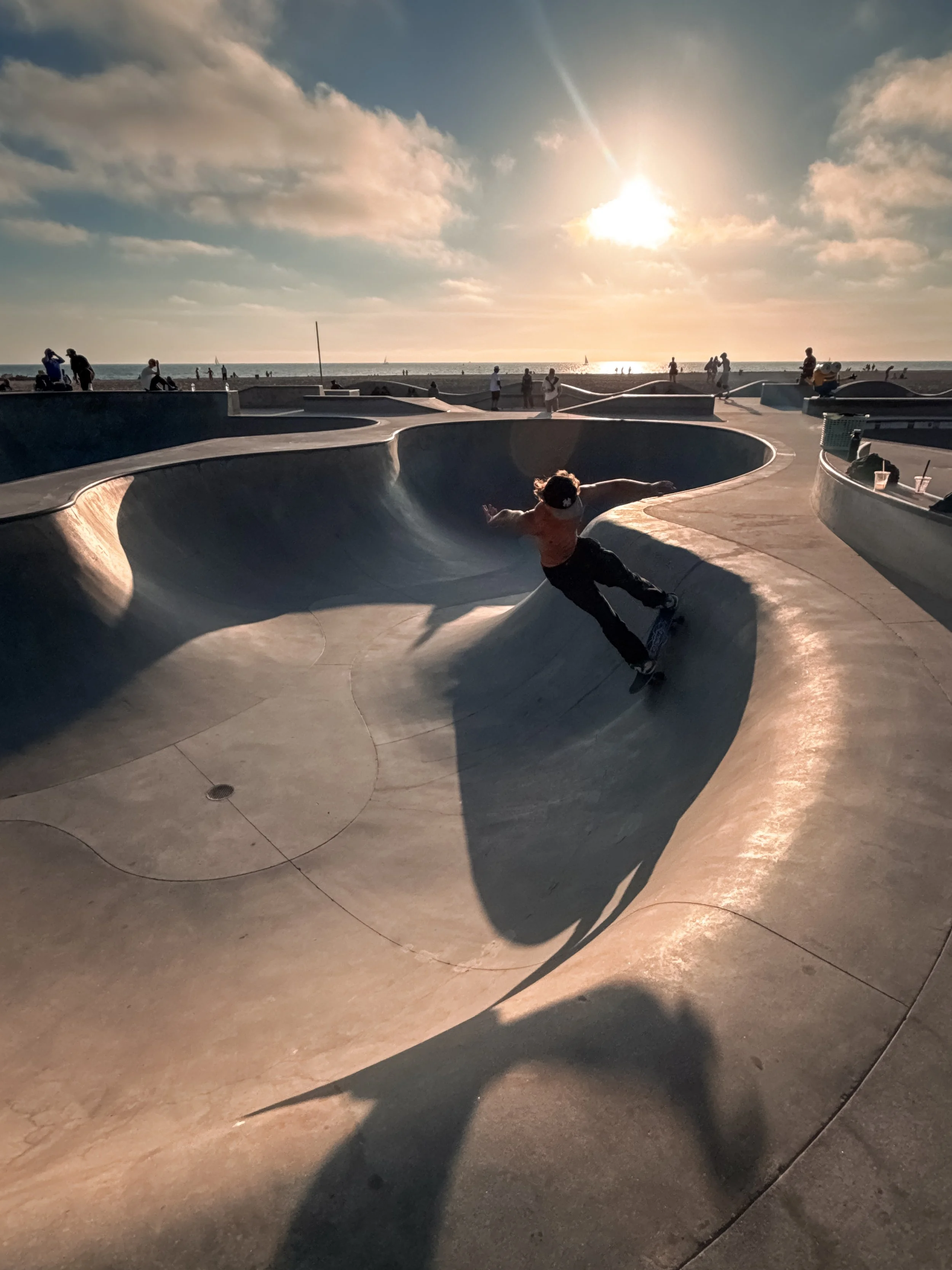 The skatepark at Venice Beach, Los Angeles, California, USA, with a person skating on a curved, smooth concrete bowl during sunset. Several people are standing and sitting around the skatepark, and the ocean is visible in the background 