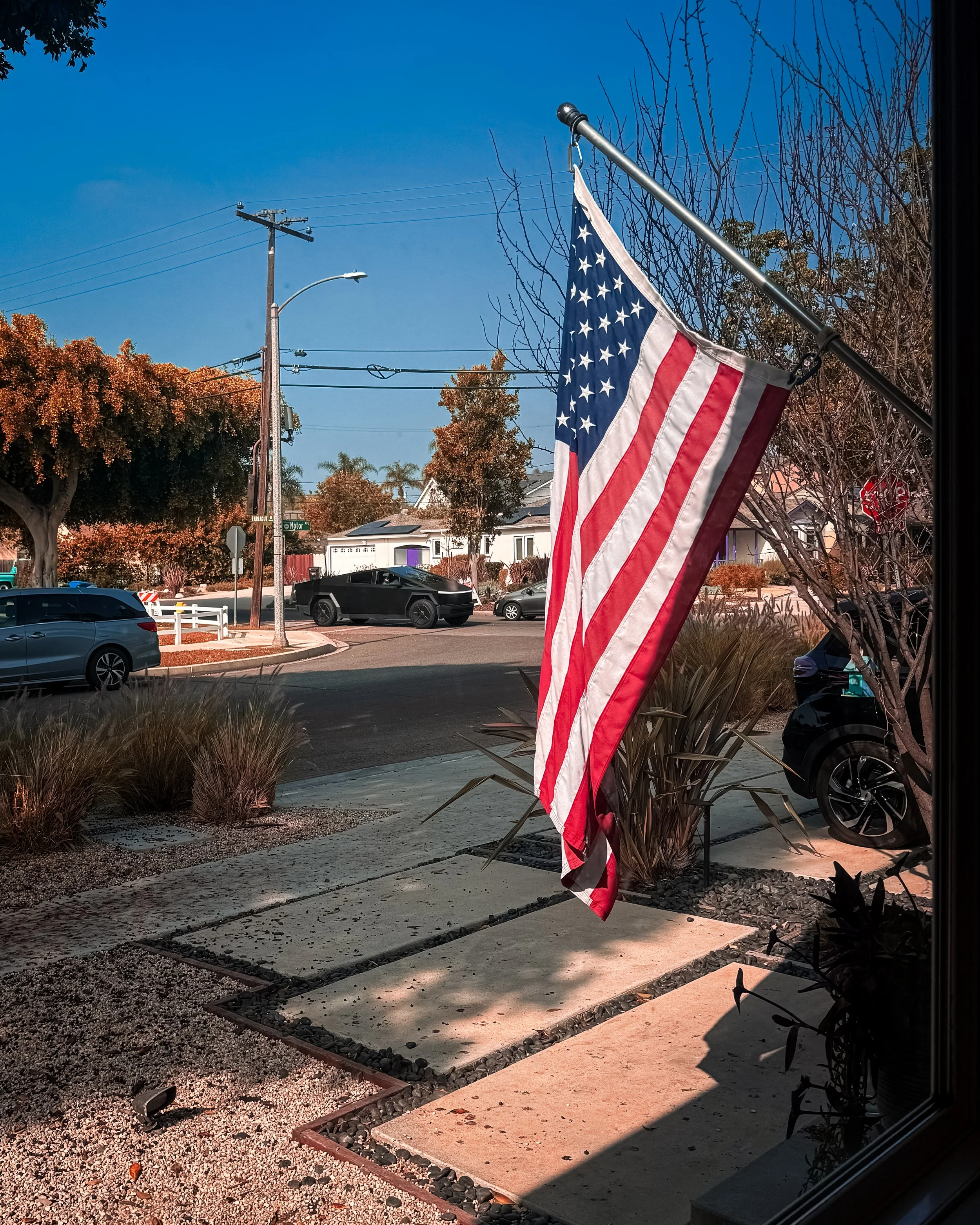 American flag hanging outside a house in the USA / America, with trees, parked cars, power lines, and a street sign in a neighbourhood under a clear blue sky. A Tesla truck can be seen on the street.