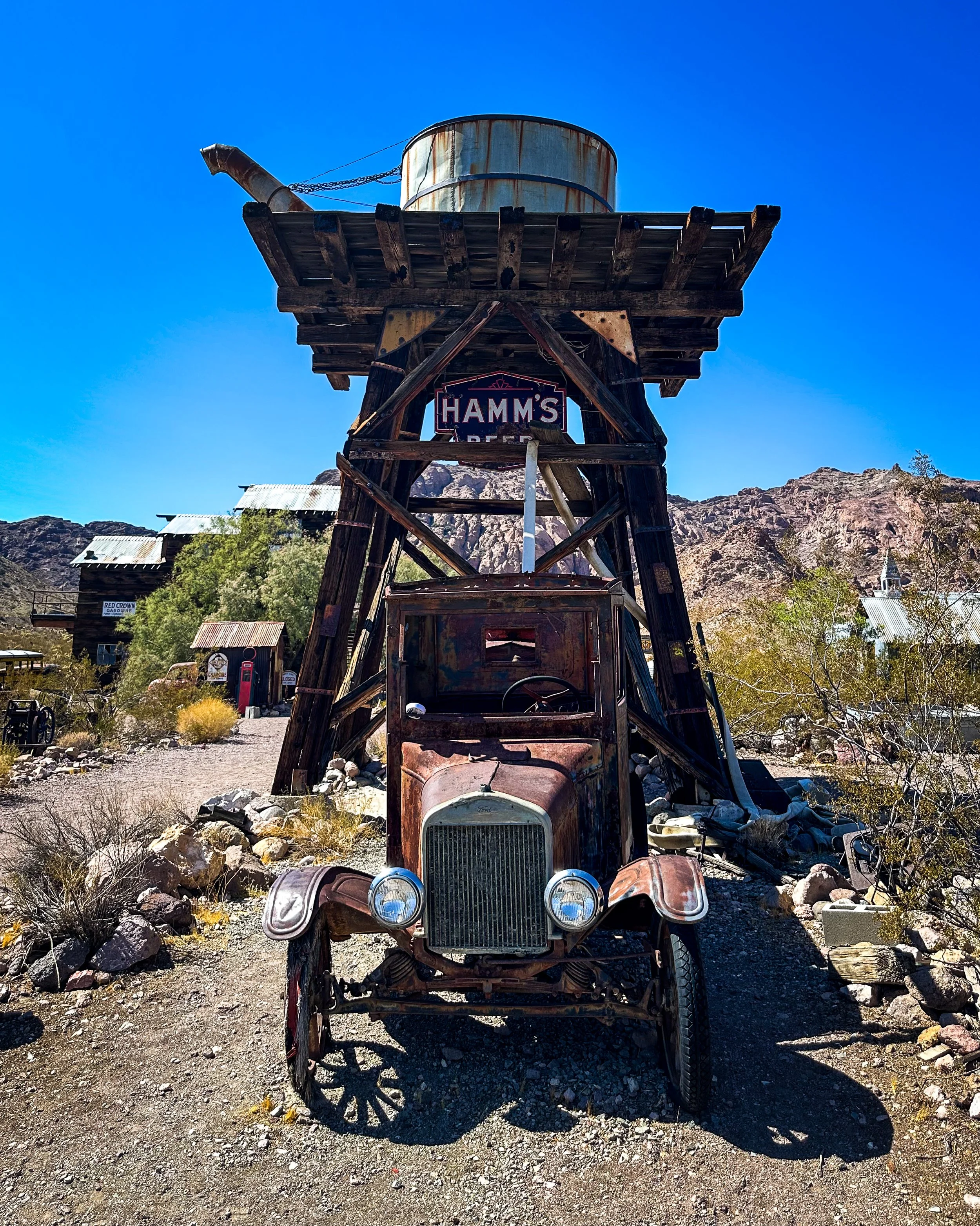 An old rusted truck under a wooden water tower with a sign reading Hamm's. The background shows a mountainous landscape with clear blue sky.