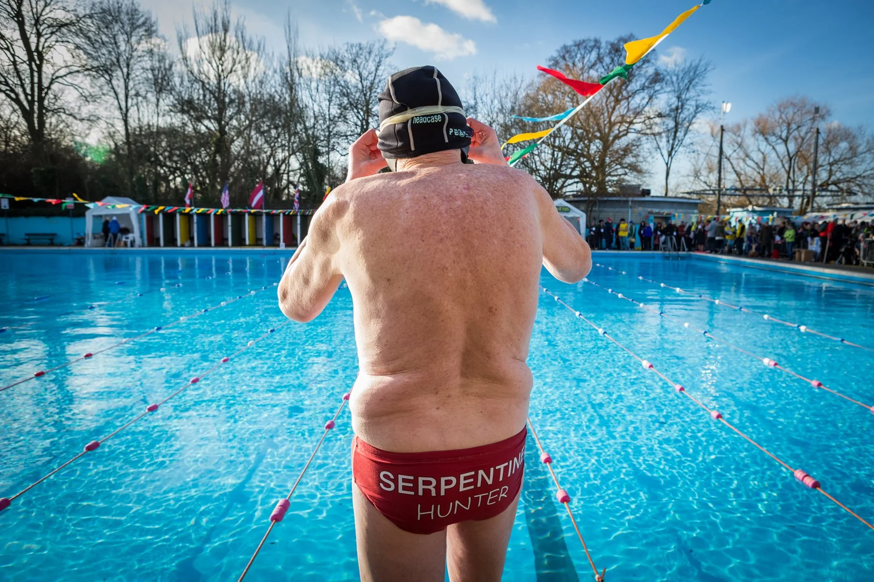 A shirtless swimmer adjusts his goggles on his head at an outdoor swimming pool during daytime. He is wearing red swim trunks with the words 'Serpentine Hunter' on the back. The pool has lane dividers and a crowd of people can be seen in the backgrou