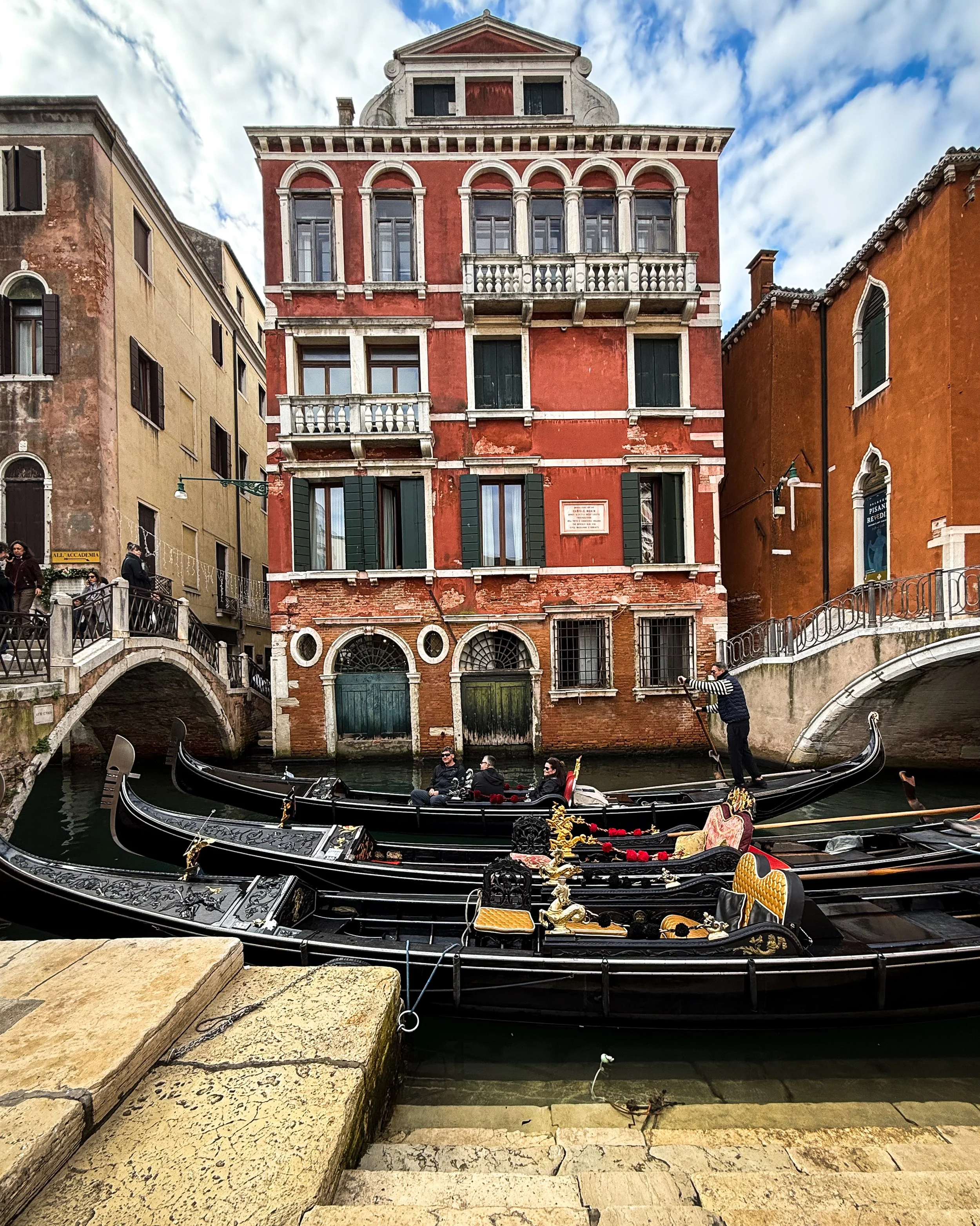 Gondolas docked along a canal in Venice, Italy, with historic buildings and a partly cloudy sky in the background.