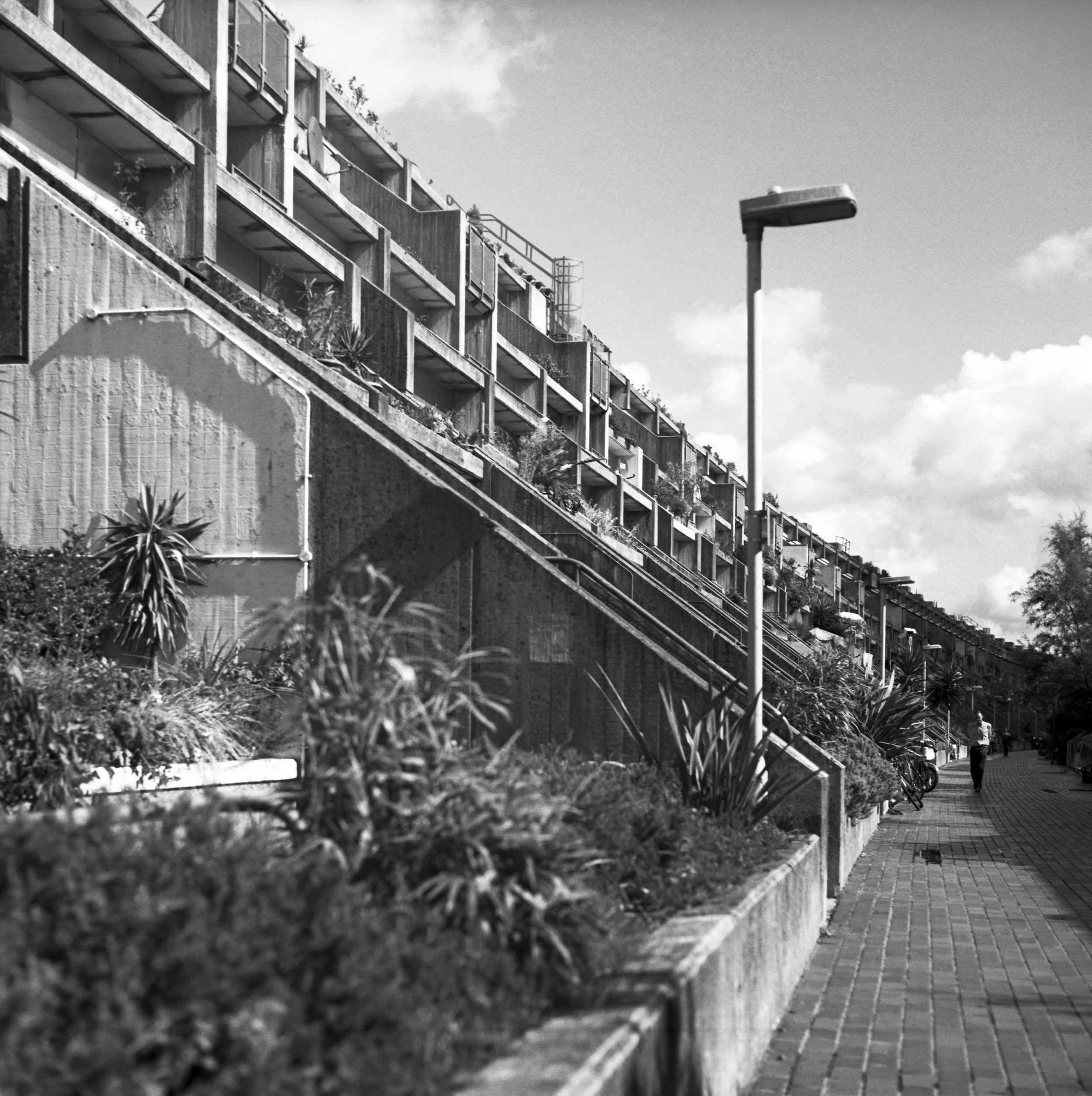 Black and white medium format photo of a multi-story residential building with balconies, some with plants, the Alexandra Estate in London. A streetlamp is in the foreground, and a man is walking along the sidewalk near some parked bicycles. 