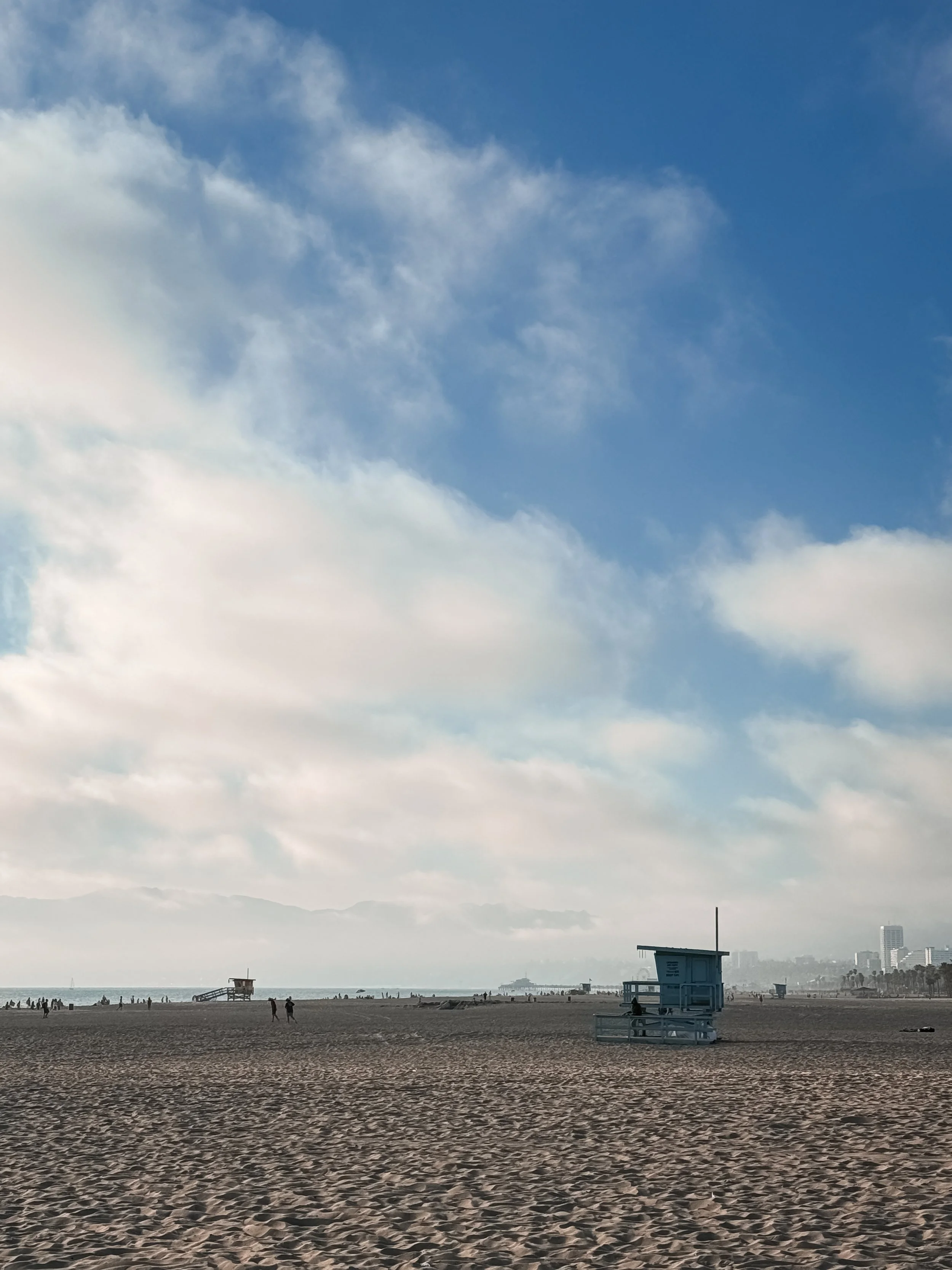 A beach scene with a sandy shore, a few people walking, lifeguard towers, and the sky filled with clouds over distant buildings.