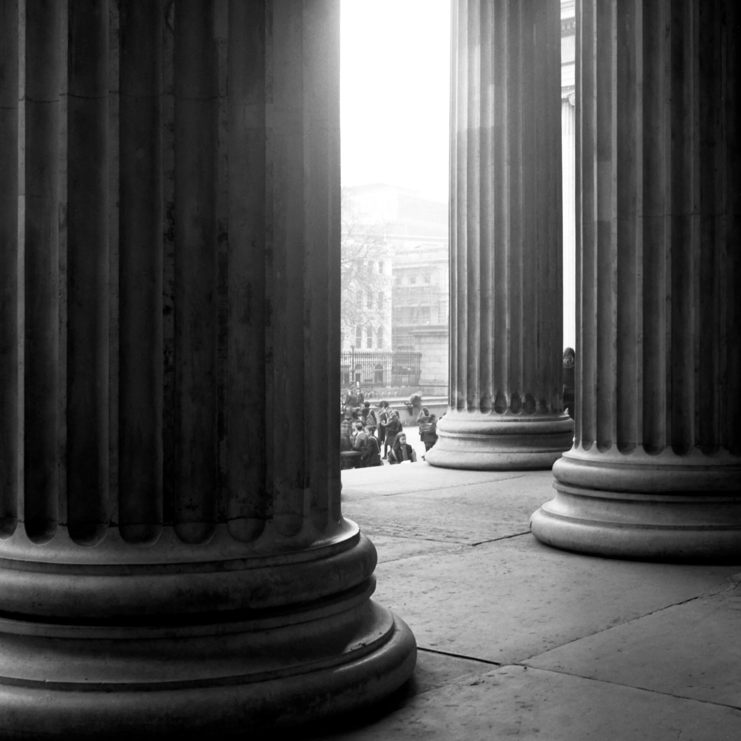 Black and white photo of large fluted columns in London, United Kingdom, and a view of a cityscape with buildings and trees beyond. 