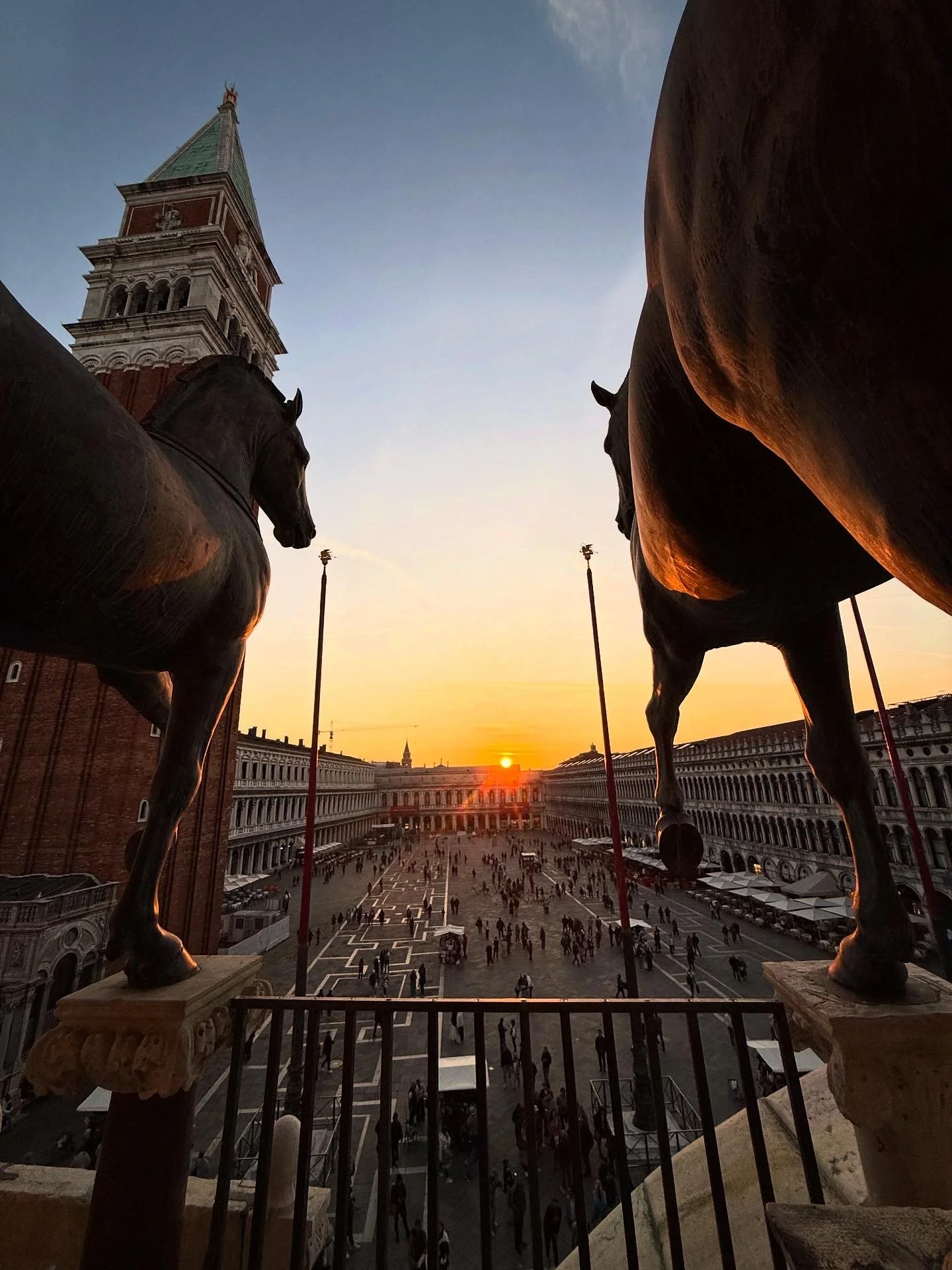 View from a balcony in Piazza San Marco, Venice, Italy, showing a sunset, the bell tower, and statues of horses silhouetted against the sky, with people walking below.