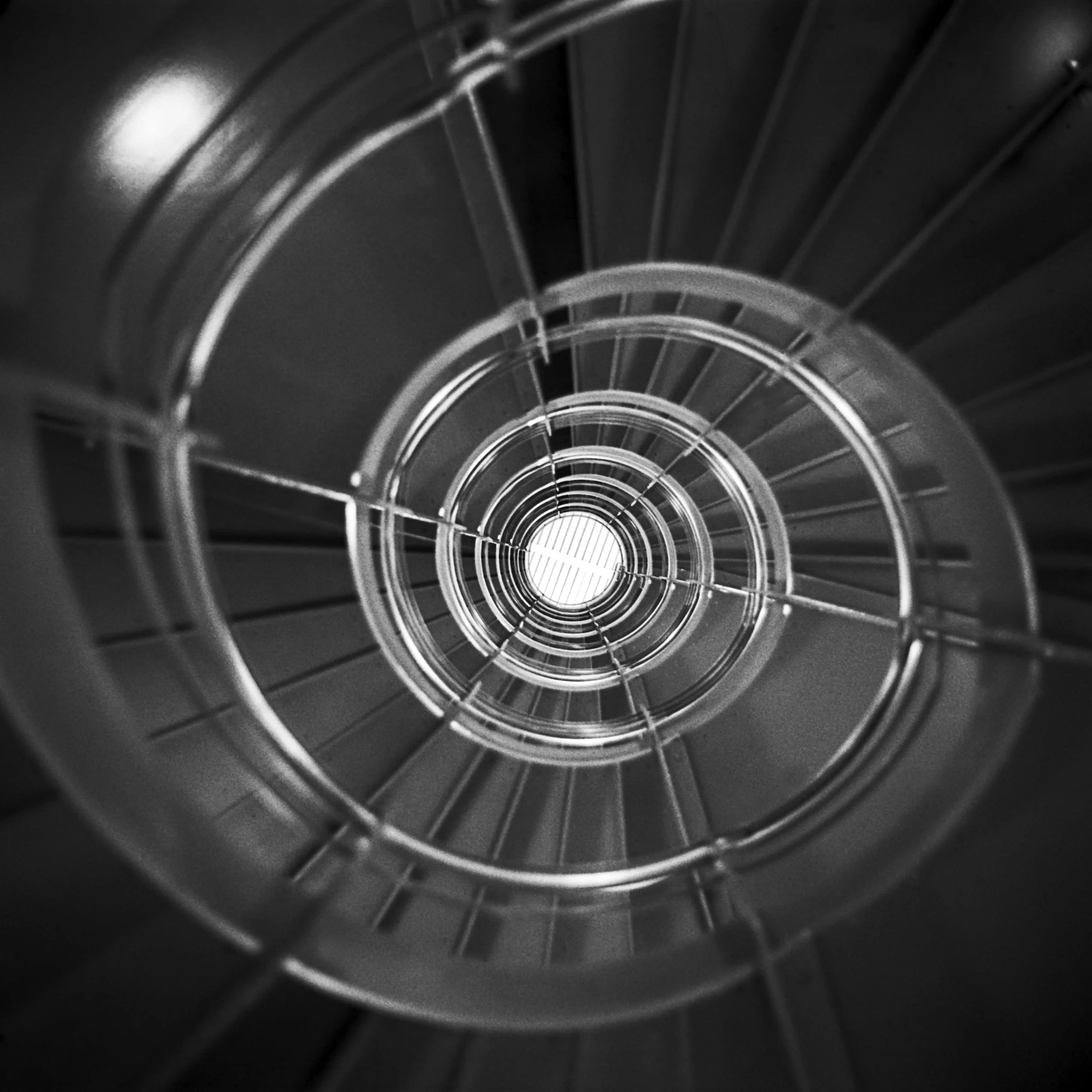 Looking down a spiral staircase with metallic railings, black steps, and a bright light at the bottom.
