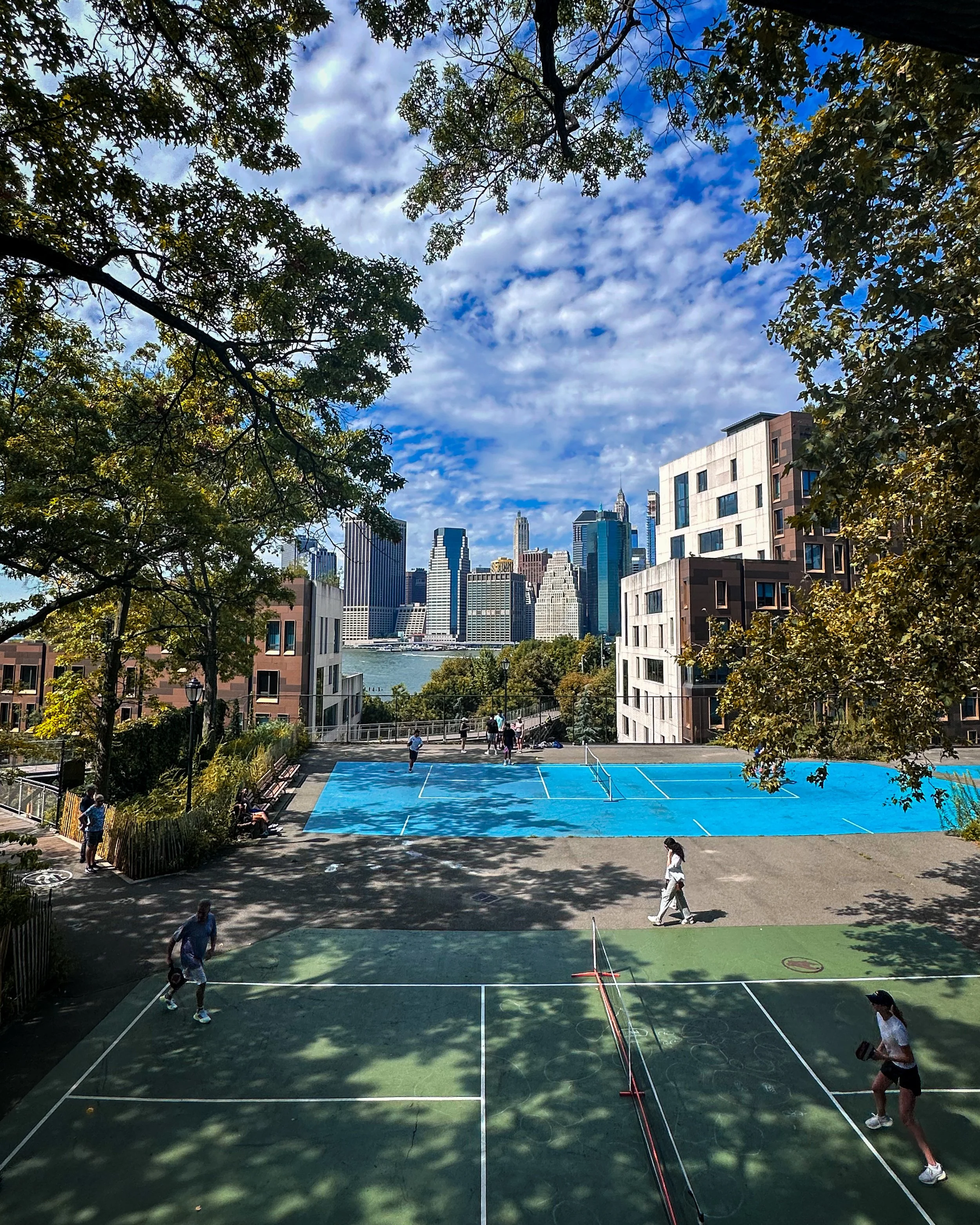 City skyline with tall buildings and a river, framed by green trees and a partly cloudy sky.