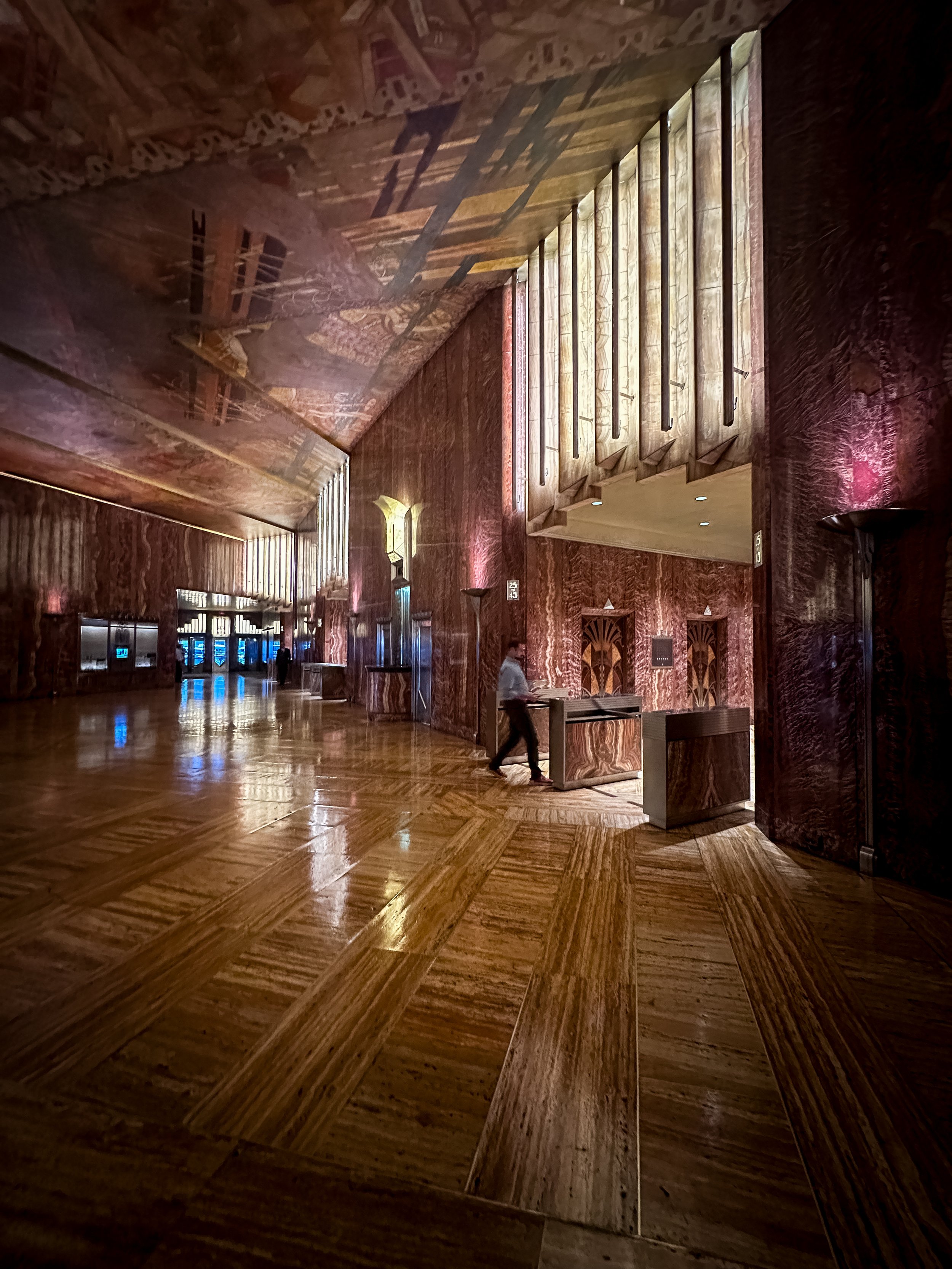 Interior of a building with dark wood paneling, a sloped ceiling, and a wood-floored lobby. There are vertical wooden blinds on a large window, and several elevator doors along the wall. A woman is walking through the space.
