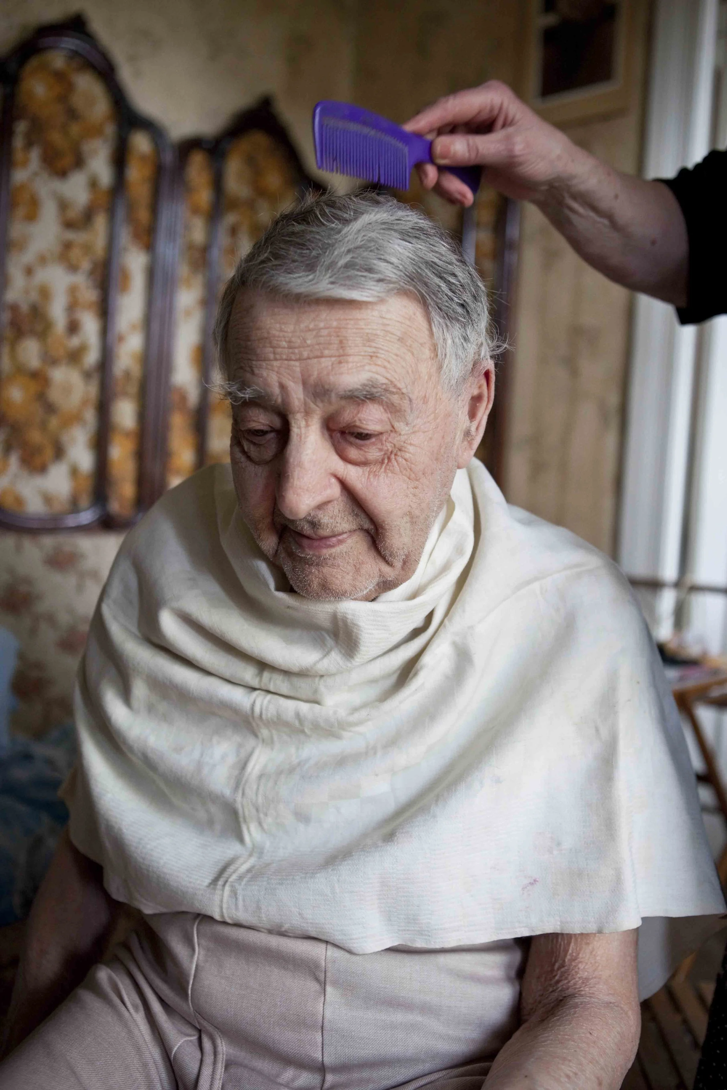 An elderly man with gray hair sitting his home in Kyiv, Ukraine, with someone combing his hair.