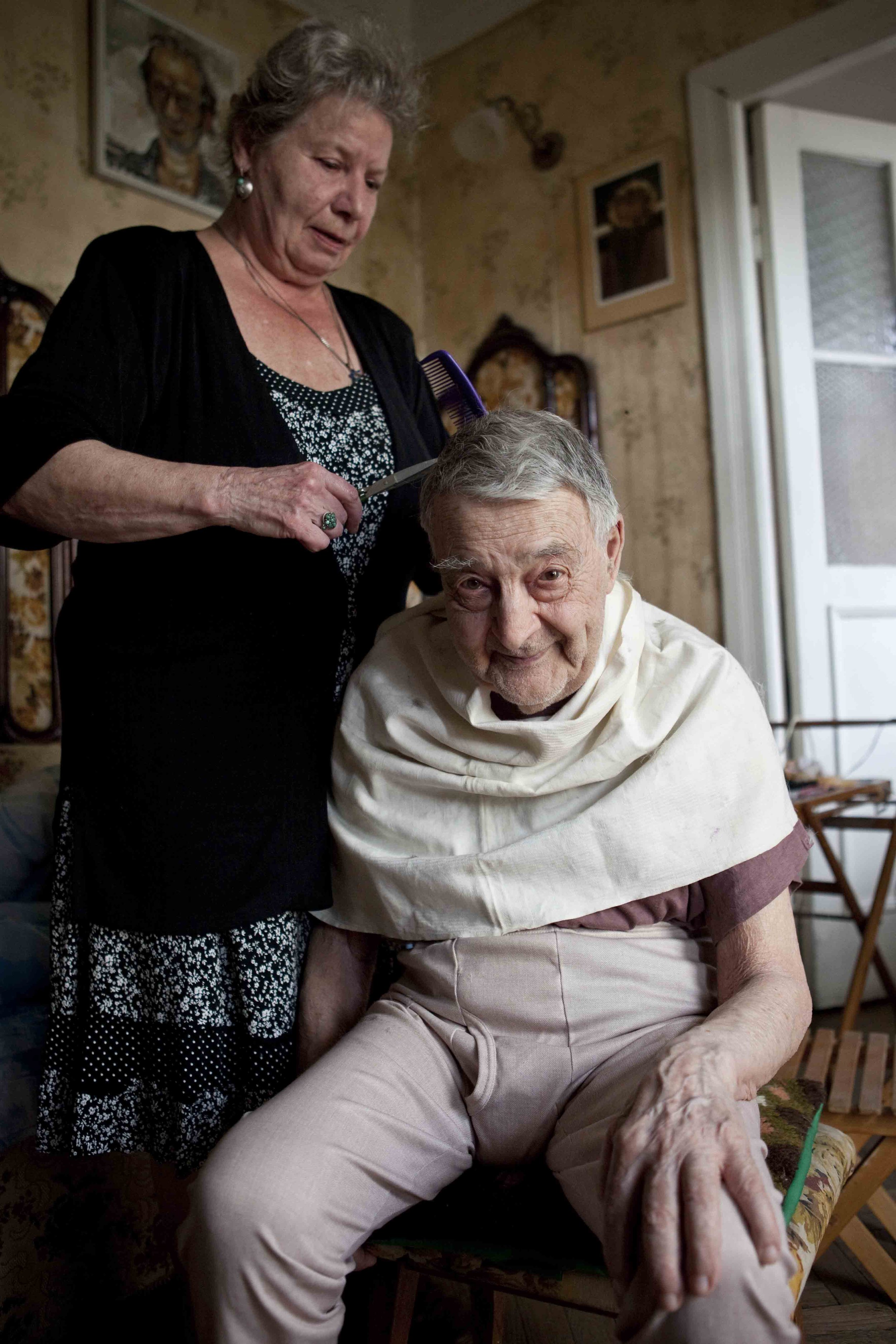 An elderly woman with short gray hair is getting a haircut from an older woman with short gray hair in a cozy room. The elderly woman is seated and smiling, wearing a beige blanket over her shoulders and beige pants. The woman giving the haircut is s