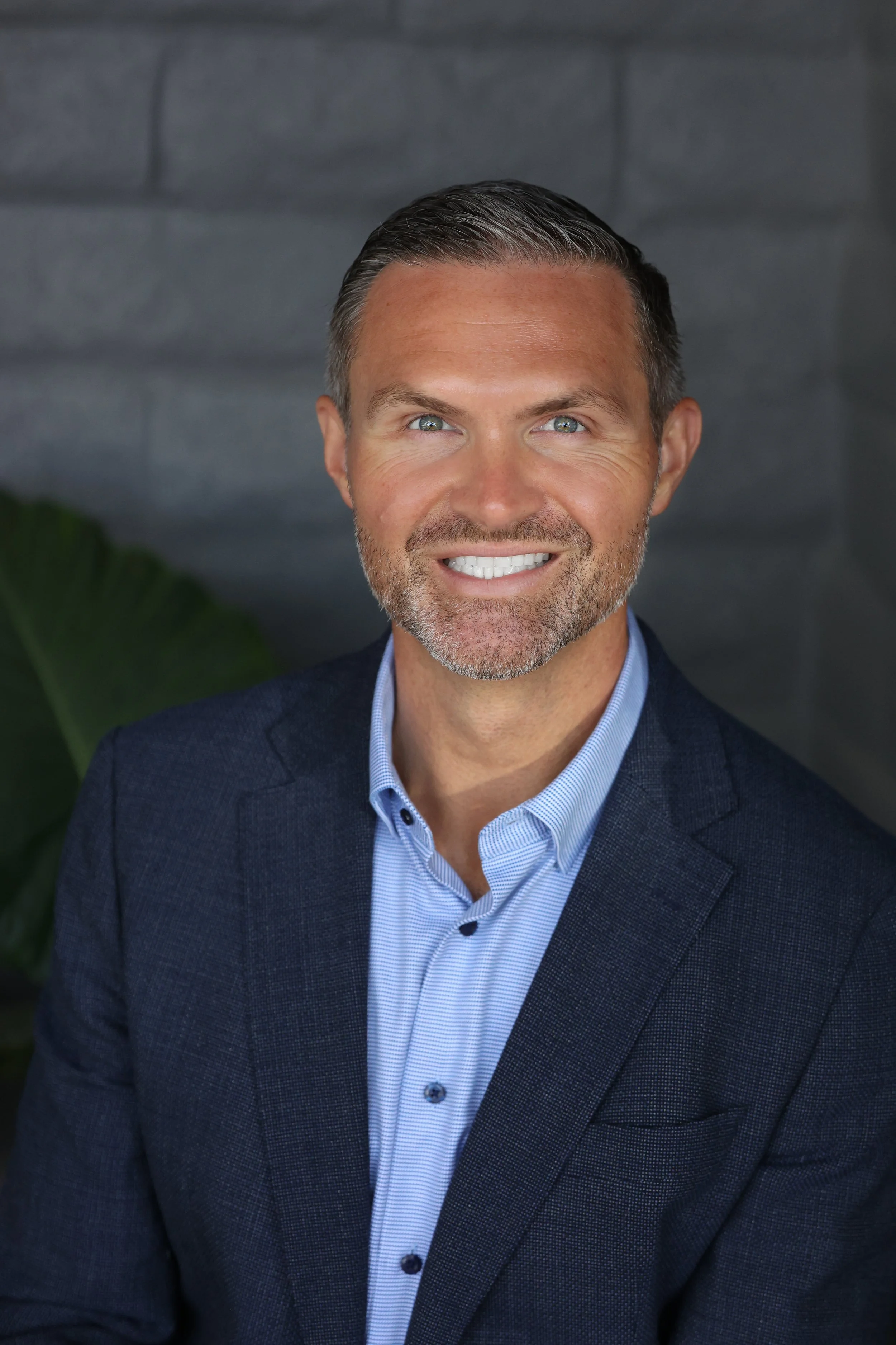 Headshot of a man with grey and dark hair, blue eyes, and a well-groomed beard, wearing a dark blue blazer and light blue shirt, smiling in front of a dark background with a large green leaf visible on the left side.