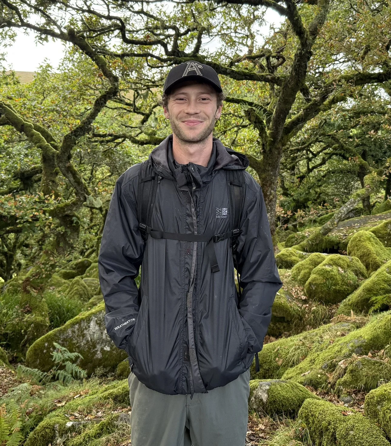 A smiling man in a black rain jacket and black baseball cap standing in a moss-covered forest with lush trees and rocks.