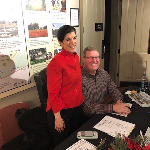 A woman in a red jacket and a man in a plaid shirt sitting at a conference table, smiling. The woman is standing next to the man, who is seated. There are papers, a phone, and a water bottle on the table.
