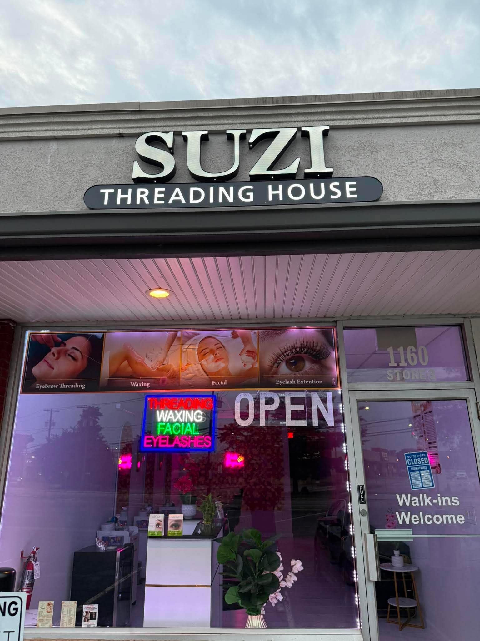 Front view of a beauty salon storefront with a sign that reads "SUZI THREADING HOUSE". The window displays images of eyelash extensions, eyebrow threading, waxing, and facial treatments. Neon signs in the window advertise services like threading, wax