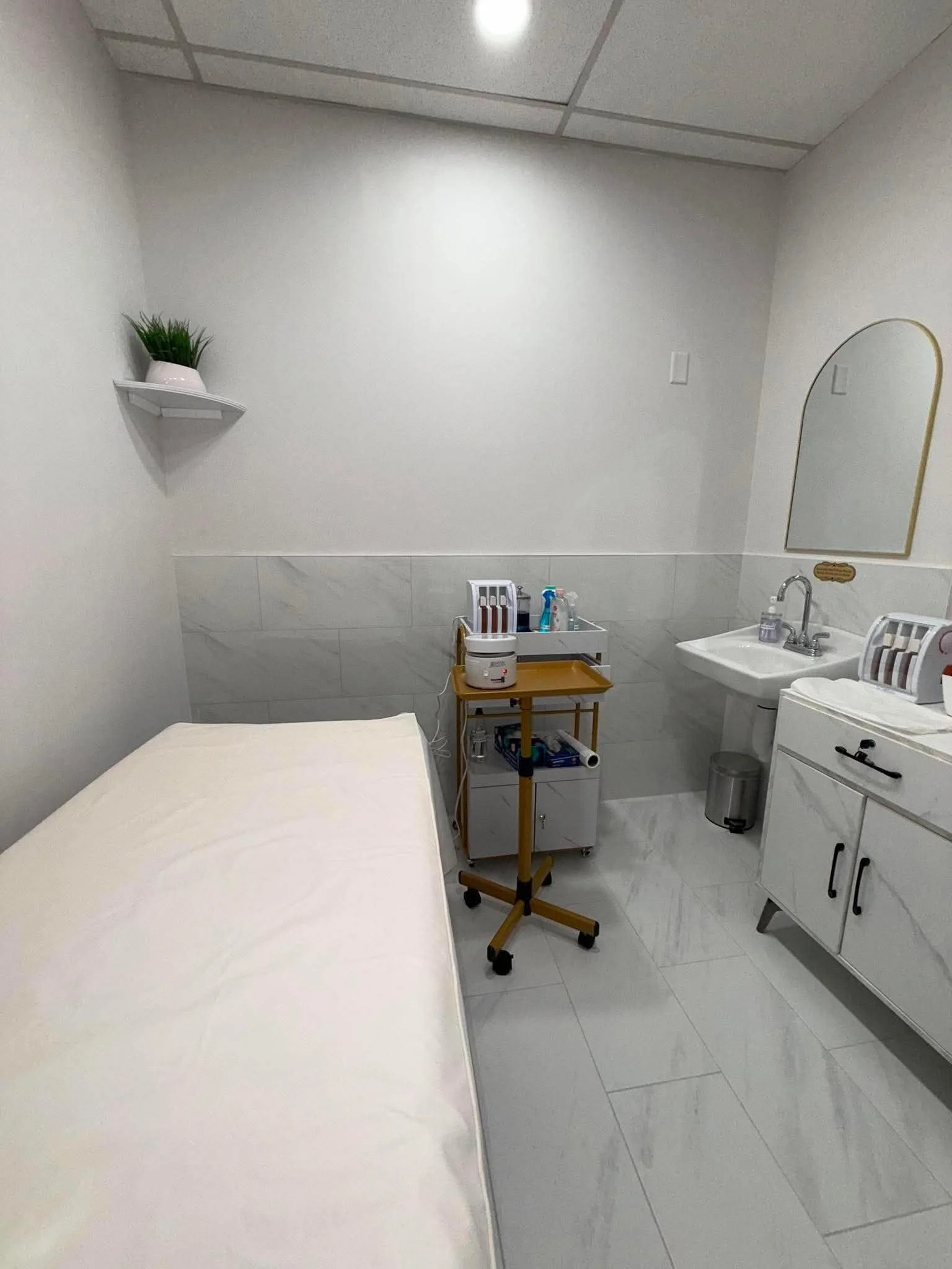 A small hospital or clinic room with a bed covered with a white sheet, a side table with medical supplies, a standing medical cart, a sink with a mirror, and a wall-mounted shelf with a plant. The room has white walls, tiled floor, and ceiling with r