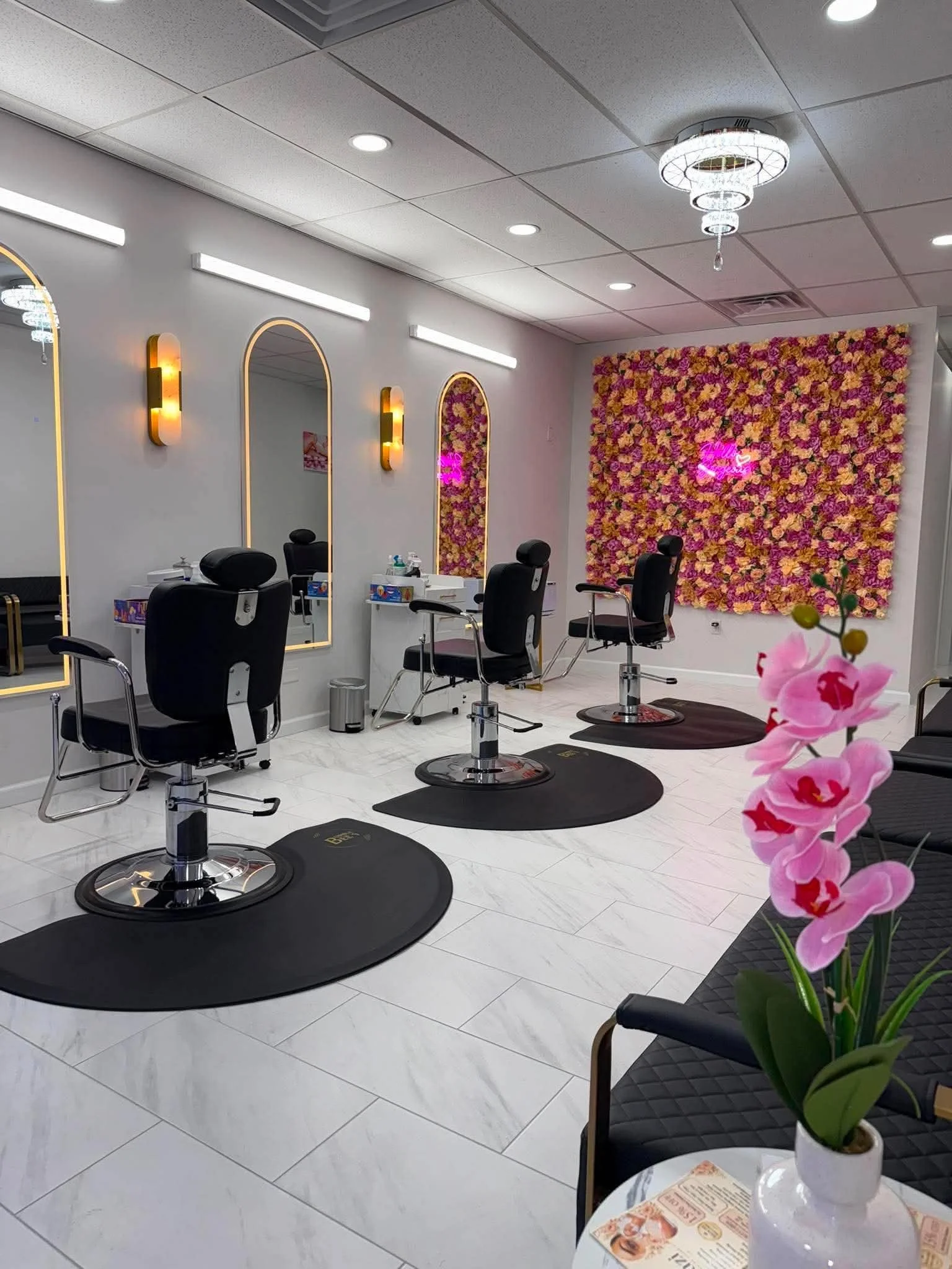 Interior of a modern hair salon with styling chairs, mirrors, and a decorative floral wall. A pink neon sign on the wall reads 'Love'. A small table with a pink orchid plant and a magazine is in the foreground.