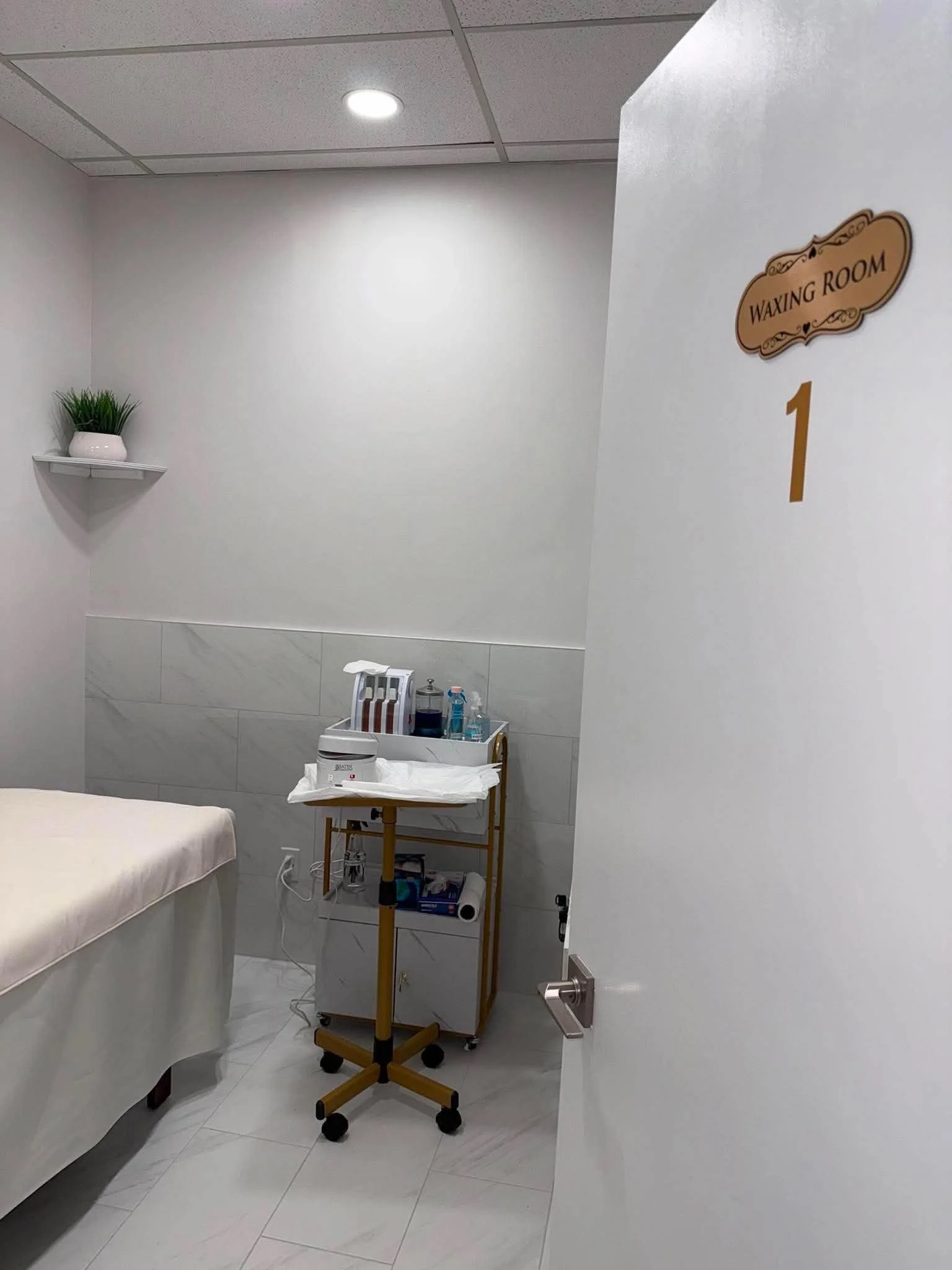 A room with a white wall and a sign that reads 'WAXING ROOM 1'. There is a small shelf with a plant on the left, a bed with a white sheet on the left side, and a medical cart with supplies in the center. The floor is tiled, and the room is lit with o