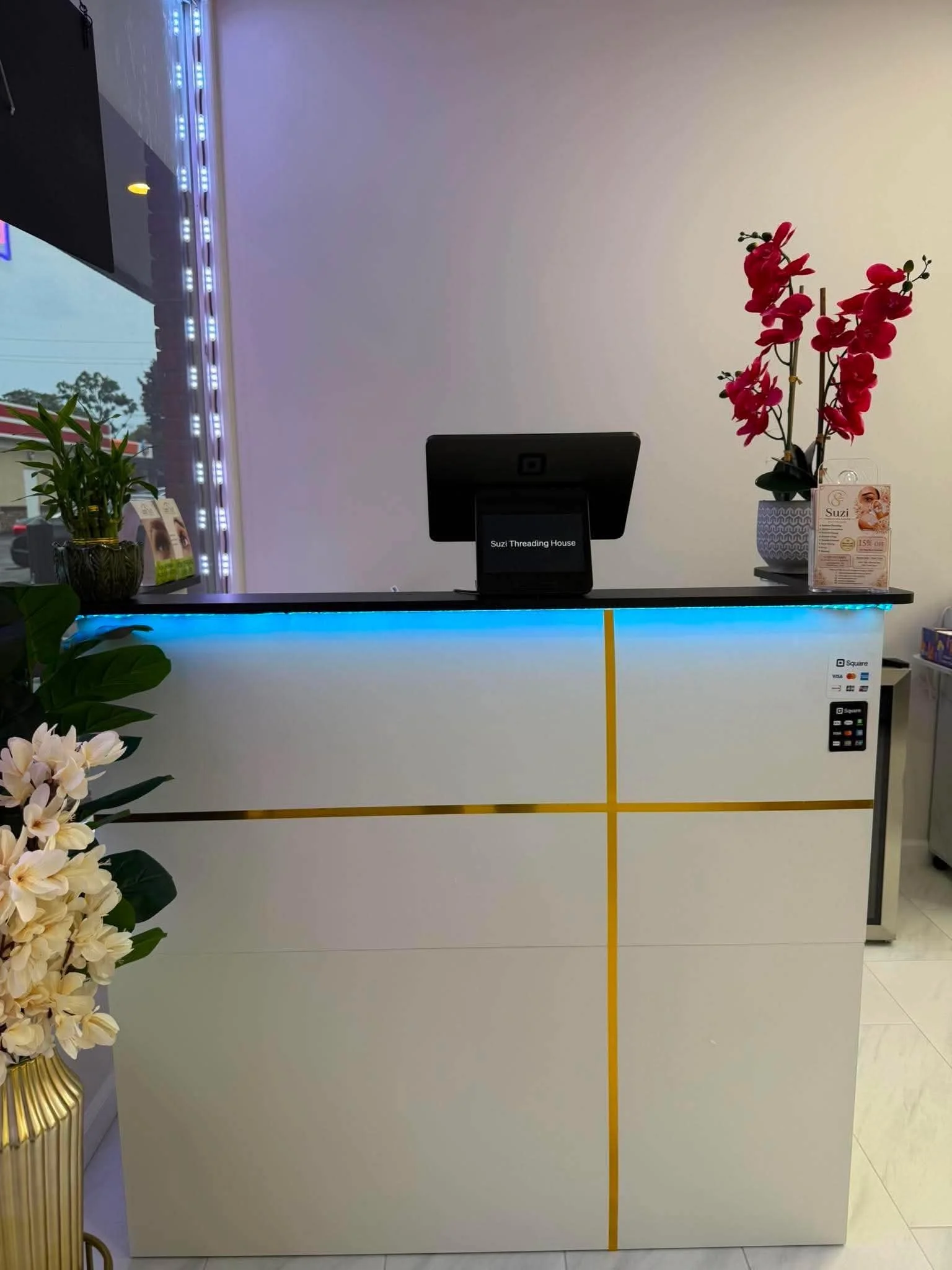 A reception counter with a black digital register, a potted pink orchid, a plant, and a promotional flyer, with a window showing a parking lot outside.