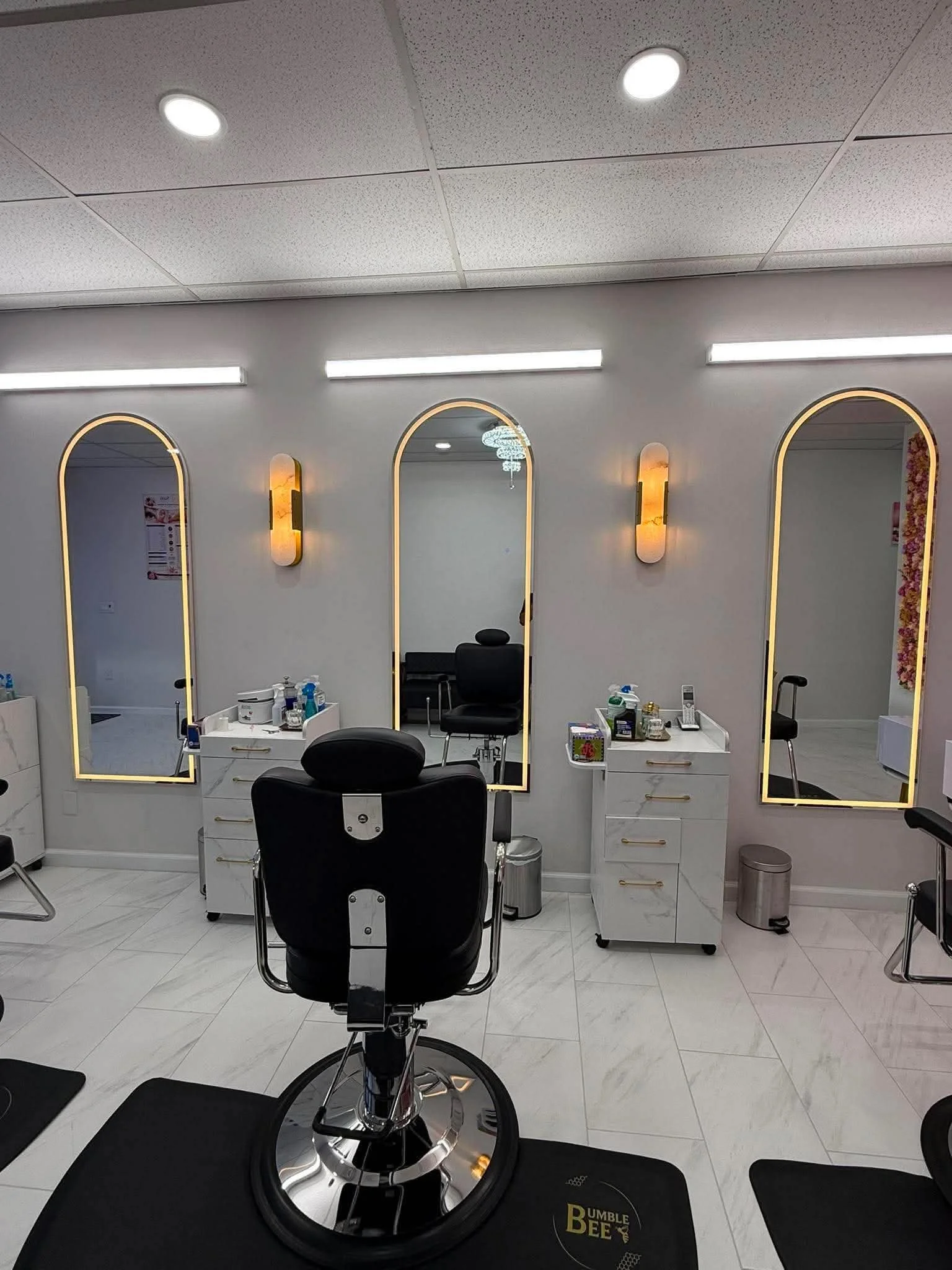 A modern barber or salon station with three mirrors, a black barber chair, and various grooming tools on white cabinets, illuminated by wall sconces and ceiling lights, with a black mat on the white tiled floor.